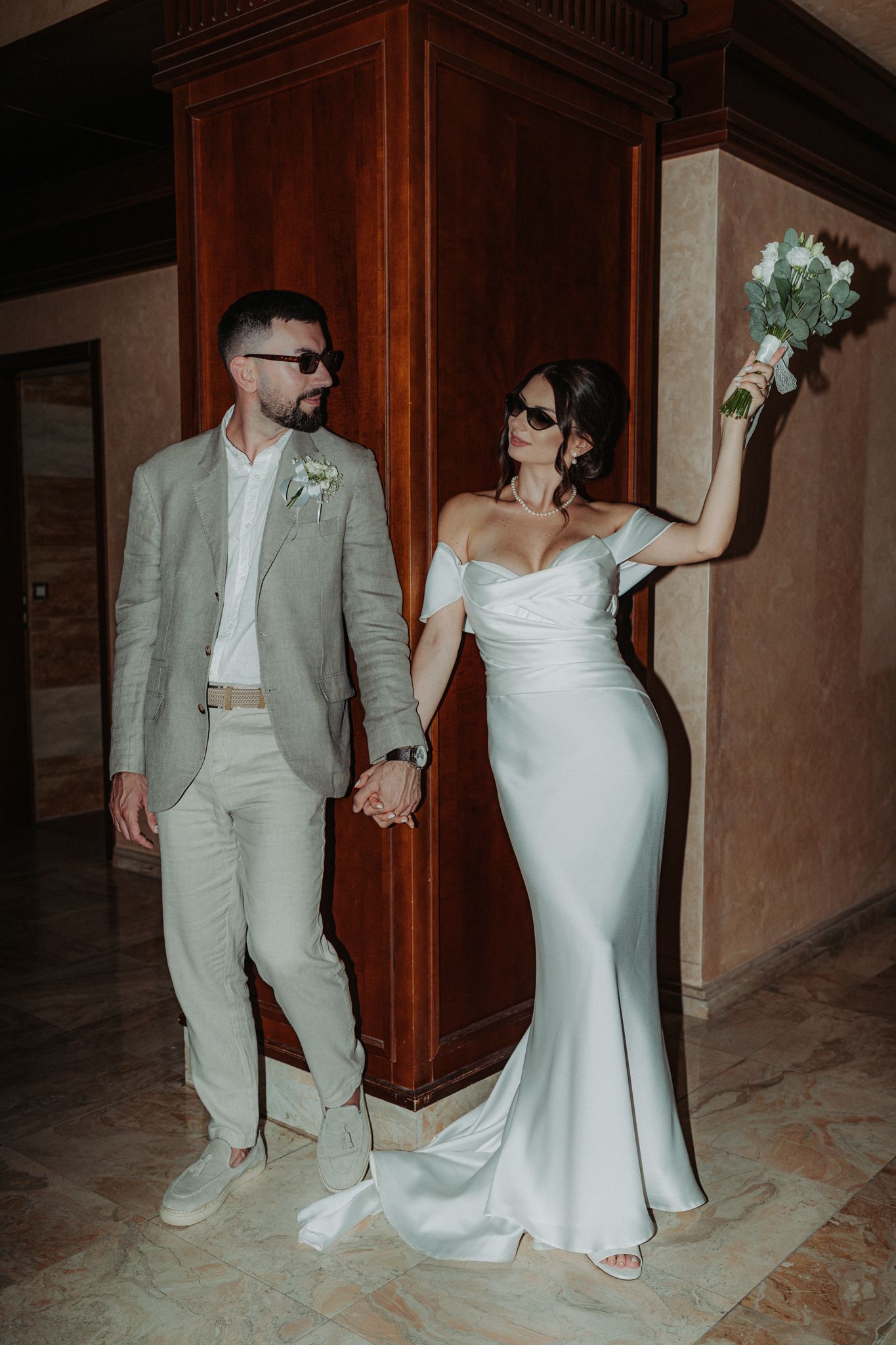 A bride in a white dress and groom in a light suit pose together in a dimly lit hallway.