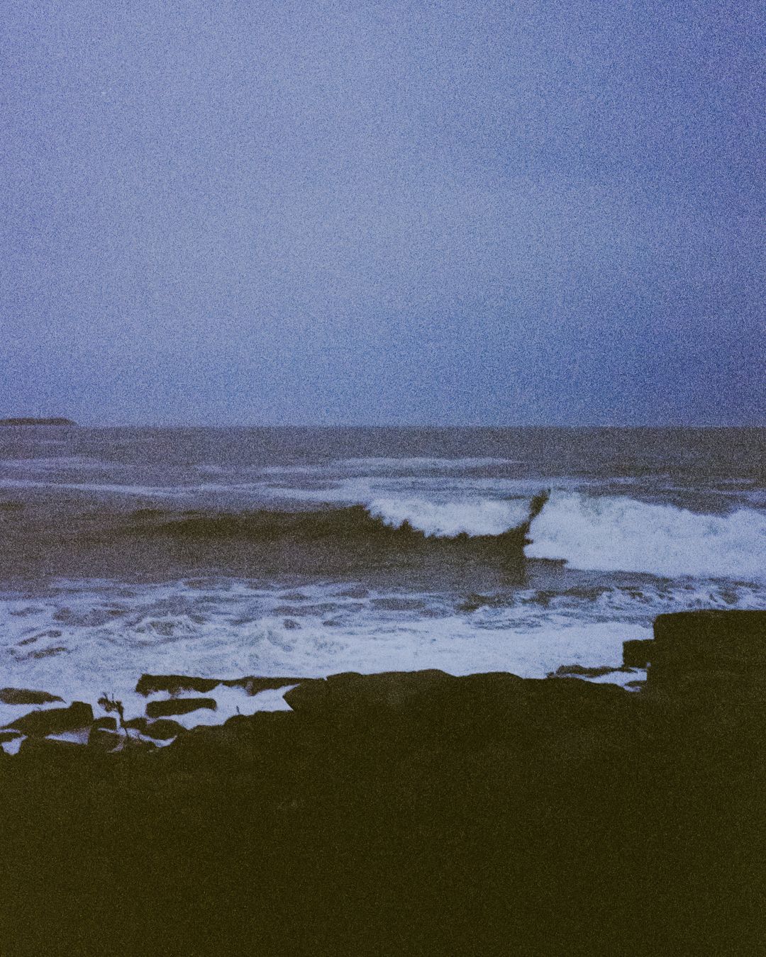 Dramatic ocean waves breaking against a dark shoreline under blue sky.