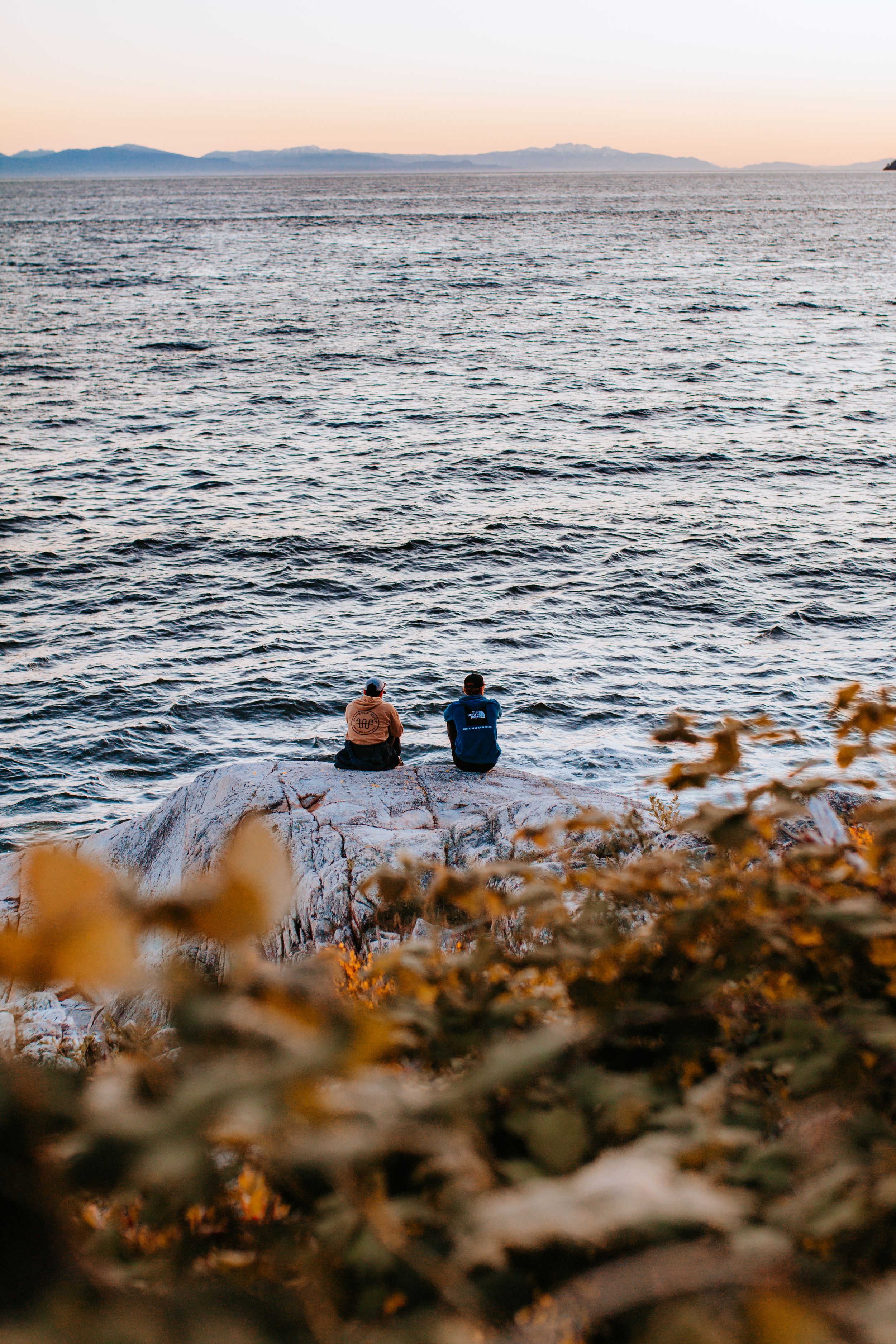 Two figures sitting on coastal rock overlooking ocean at sunset.