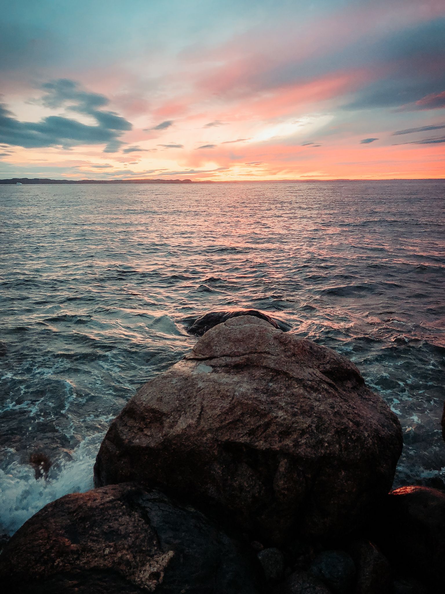 Coastal rocks at sunset with pink and orange sky reflecting on calm ocean waters.