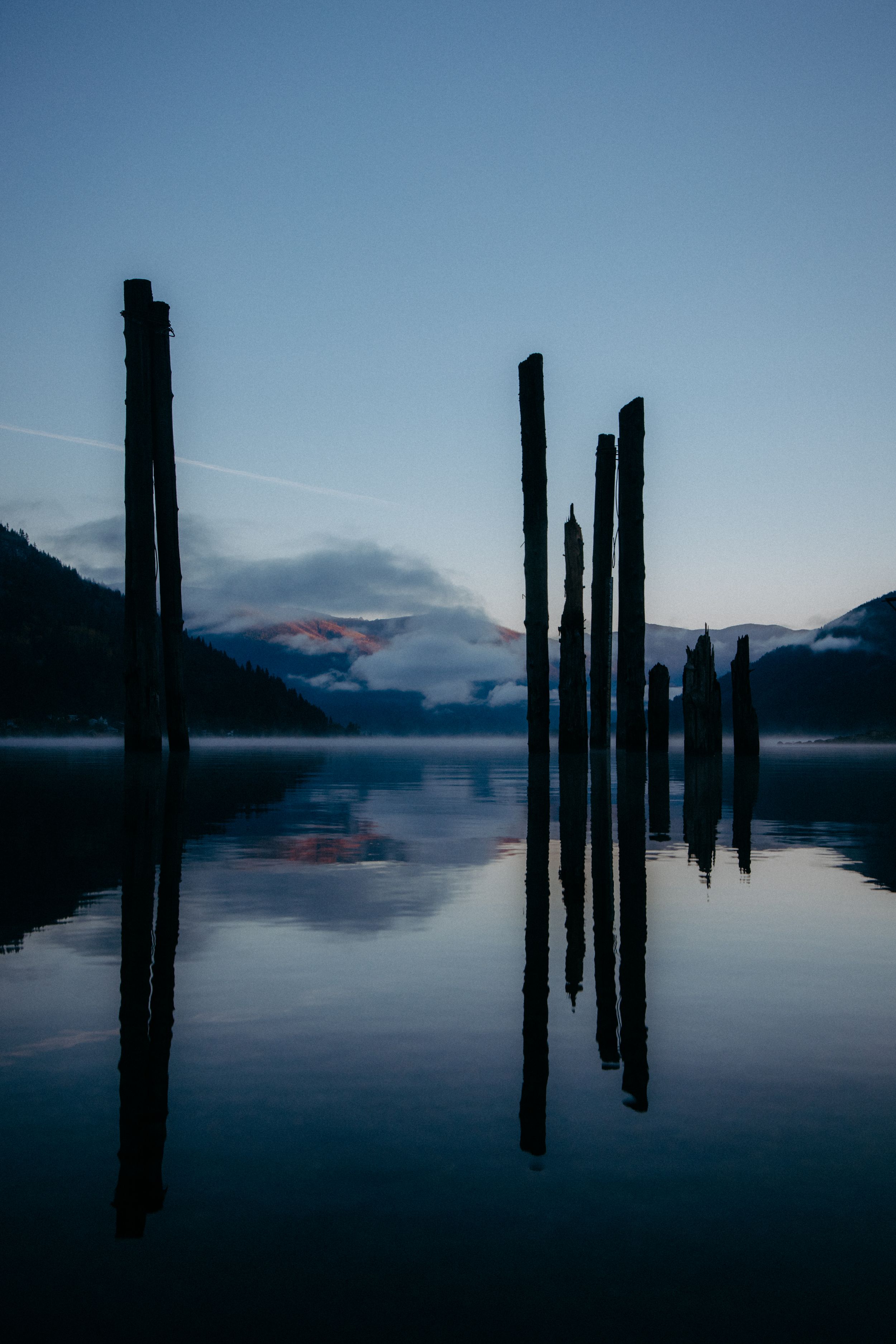 Wooden posts reflected in calm lake water at twilight with mountains in background.