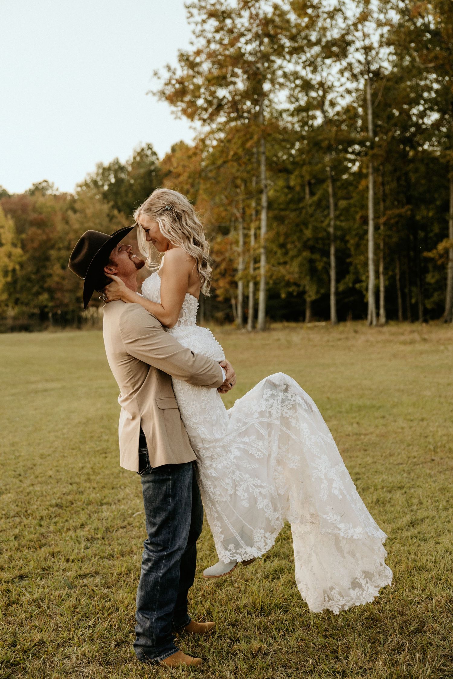 Groom in western wear lifts bride in lace dress during sunset photo session.