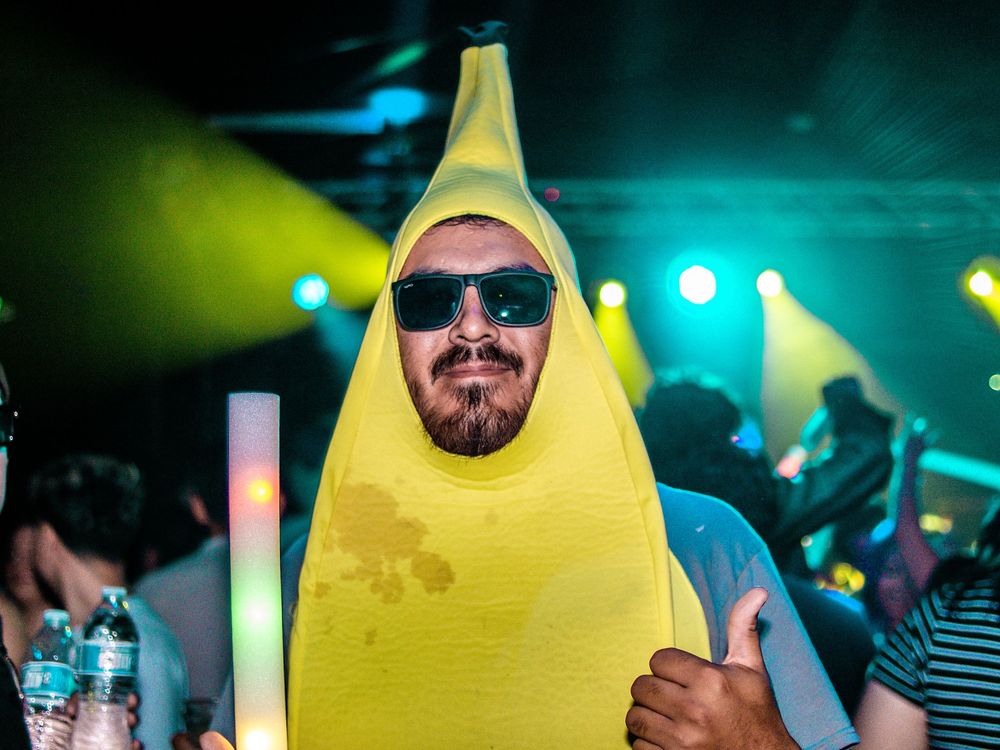 Man wearing a yellow banana costume with sunglasses giving thumbs up at a nightclub party.