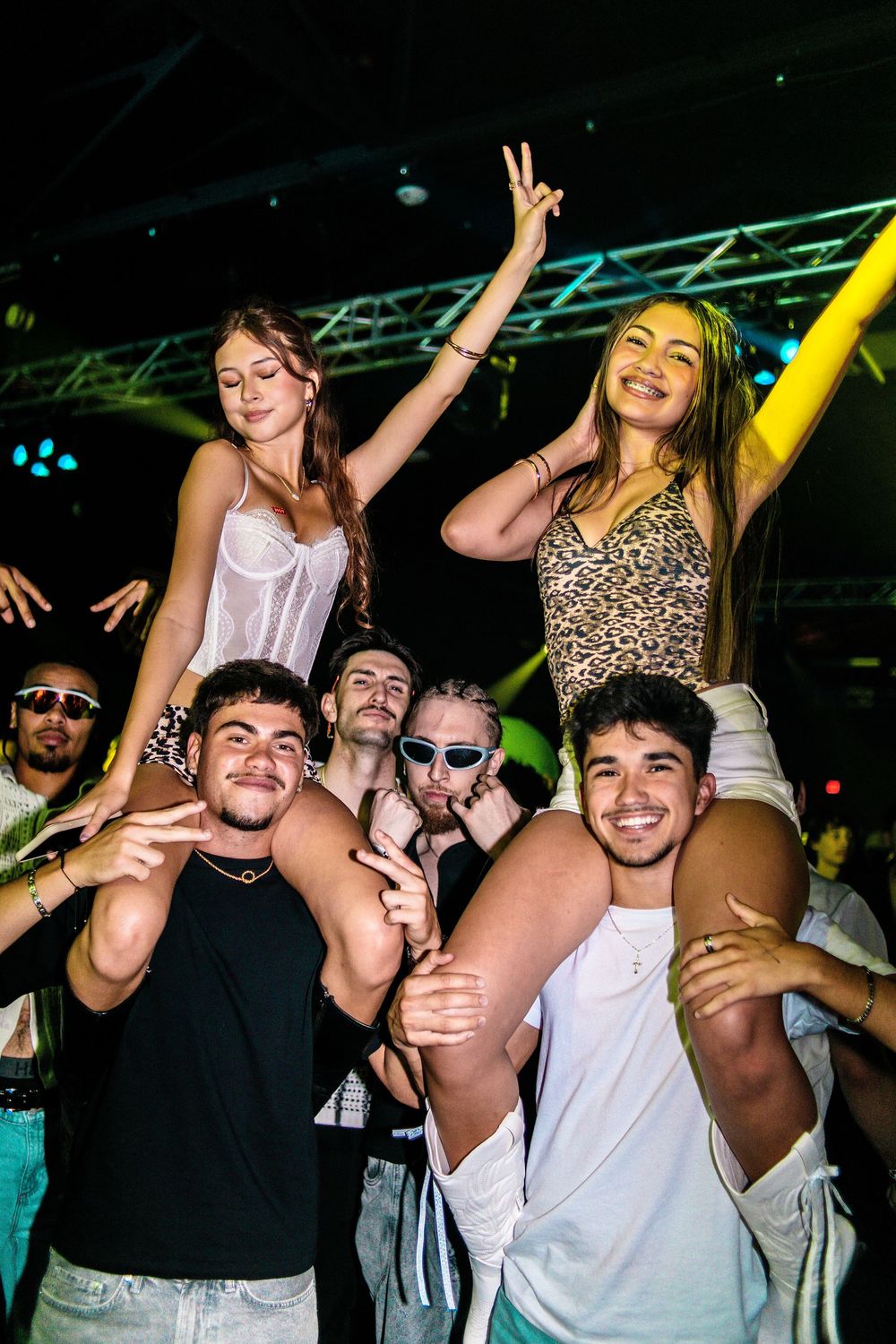 Group of young friends celebrating at a nightclub, two women sitting on shoulders dancing.