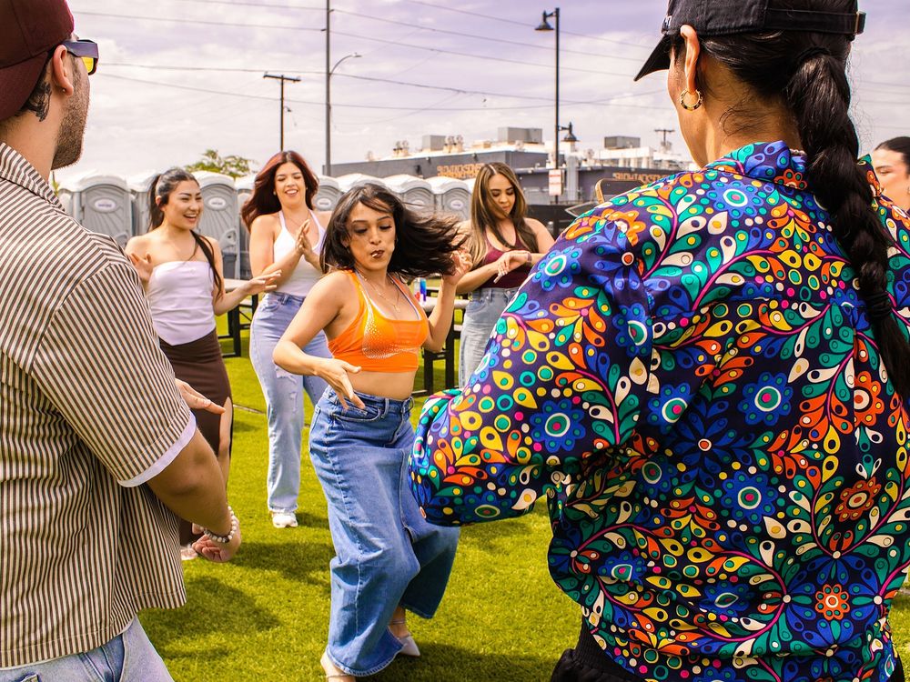 Group of friends dancing outdoors at a festival on green grass, wearing colorful summer outfits.