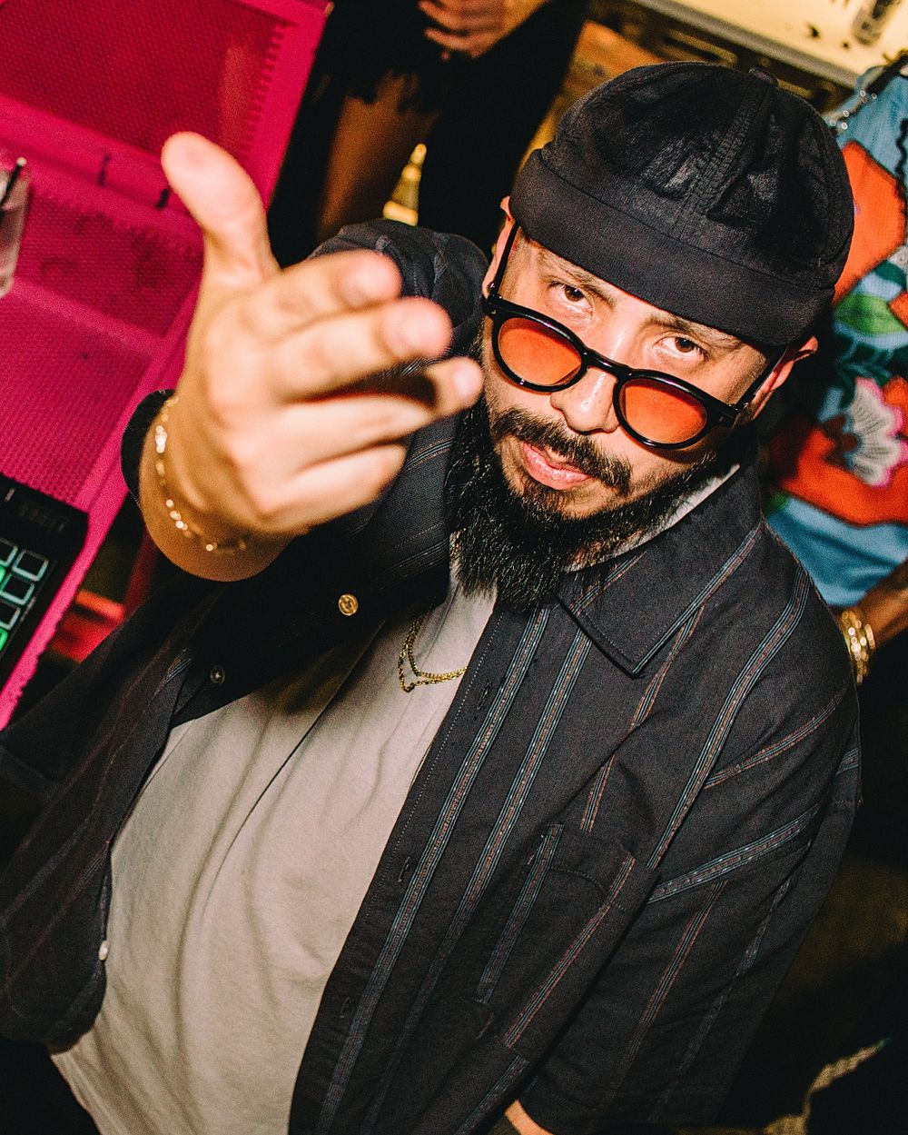 A bearded man wearing orange-tinted round sunglasses and a black cap reaches toward the camera at a vibrant event.