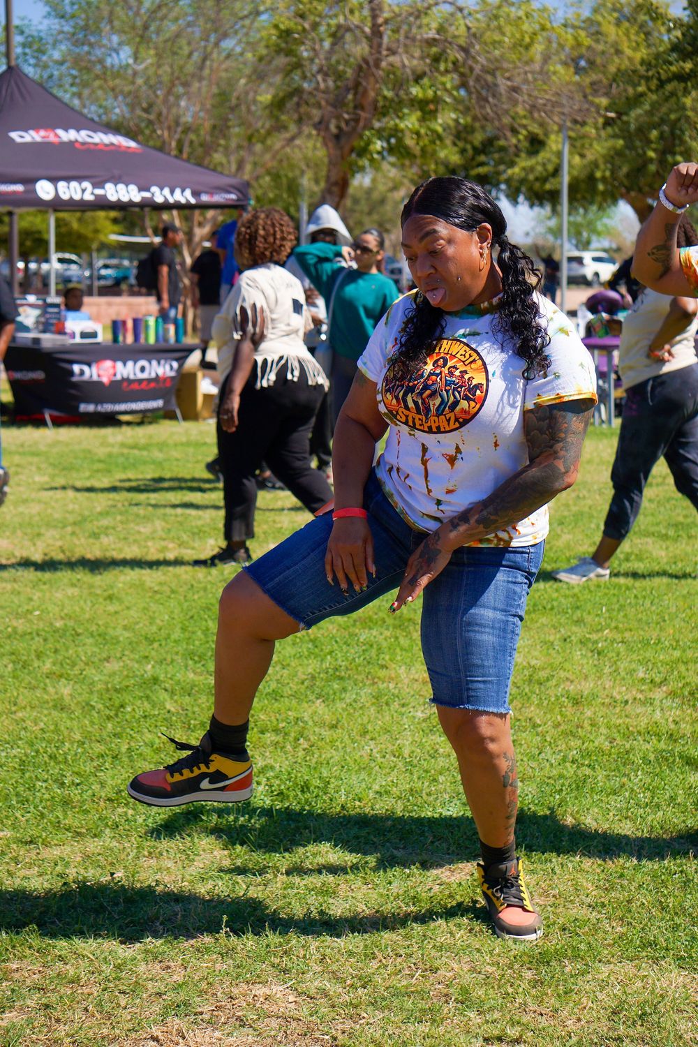 A group of dancers in casual attire practice synchronized dance moves on a grassy lawn during a sunny day.