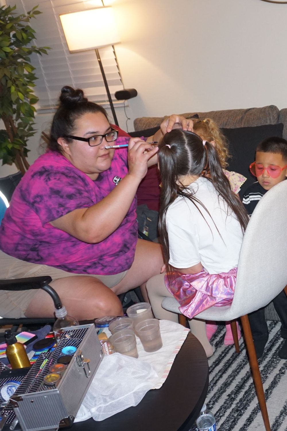 Face painting artist in pink tie-dye shirt applies colorful design at indoor party.