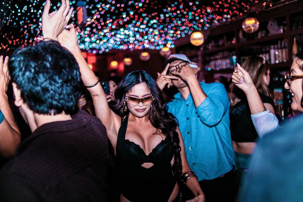 People dancing and celebrating in a lively bar with colorful bokeh lights in the background.