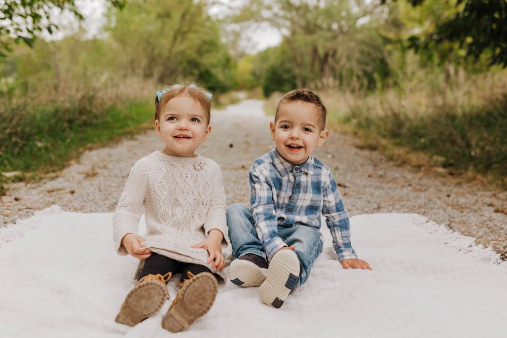 Two young children sit together and laugh while playing on a gravel path surrounded by greenery.