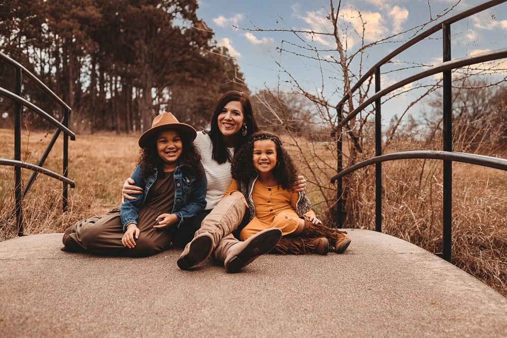 Three smiling friends sit together on a wooden bridge surrounded by autumn foliage.