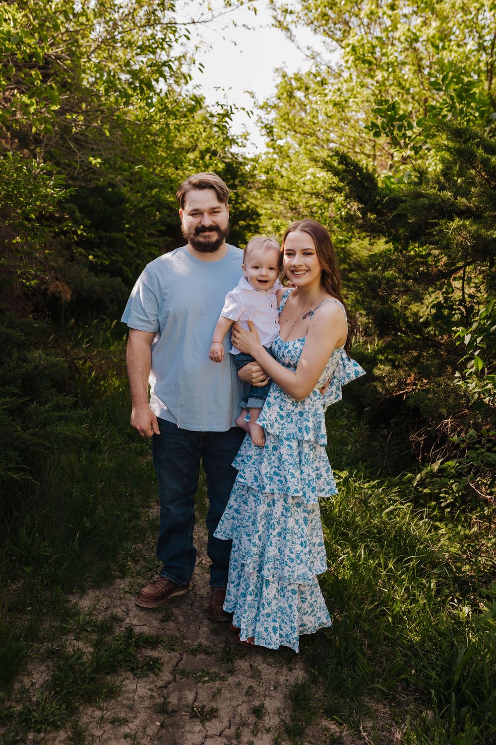 A family in casual attire poses together against a lush green outdoor backdrop during golden hour.
