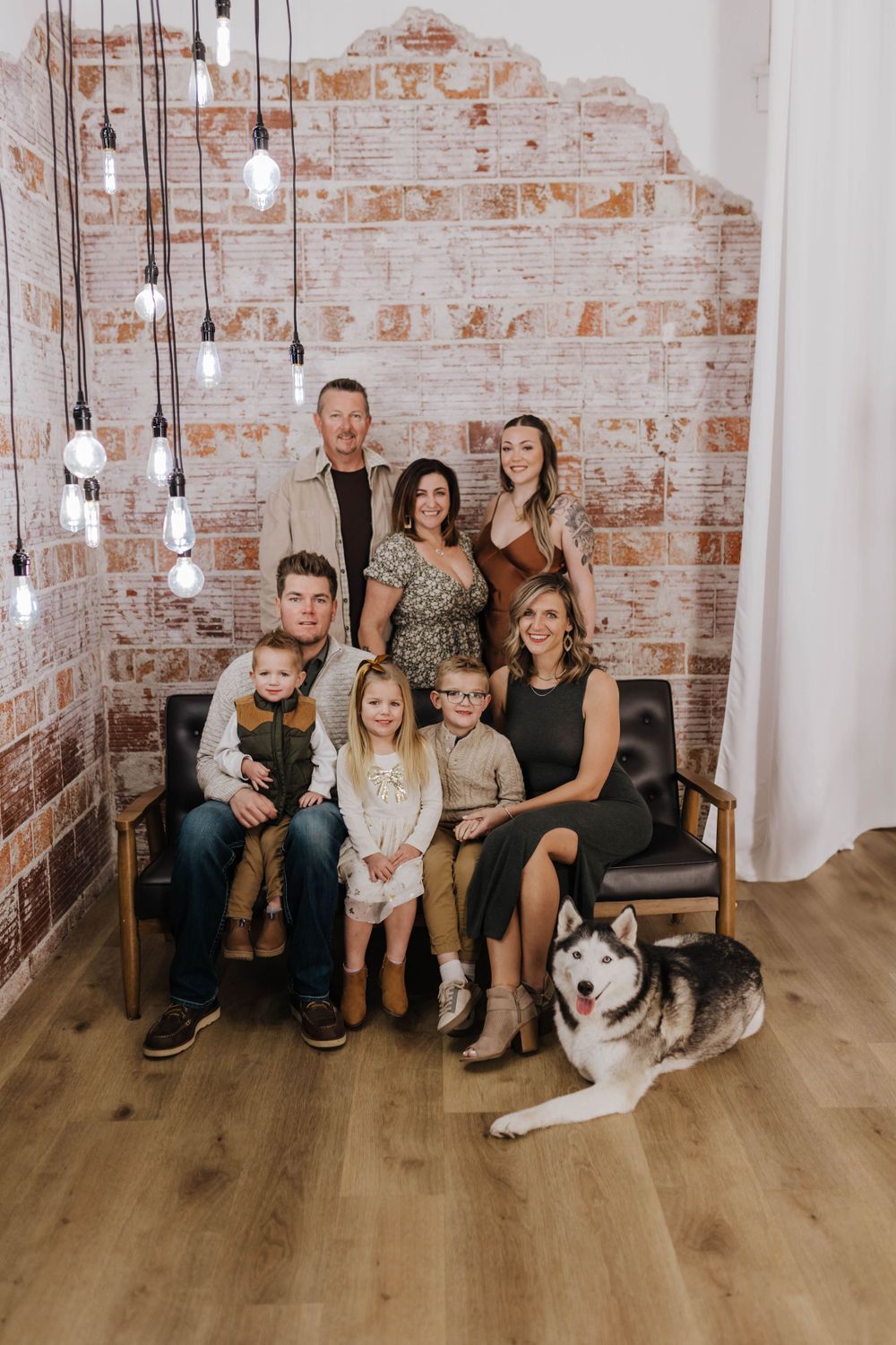 A large family portrait in a rustic room with exposed brick walls, hanging lights and a husky dog.