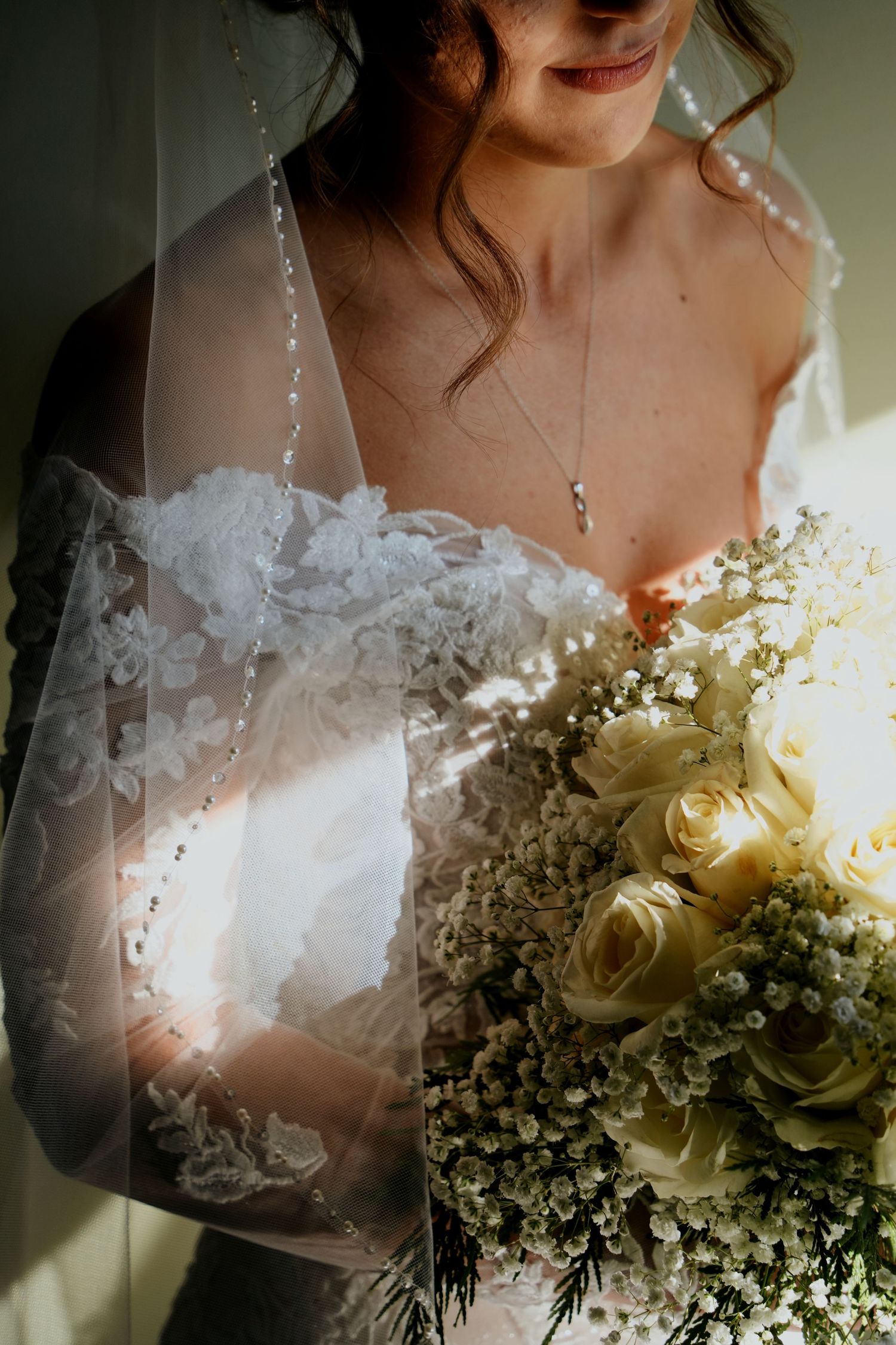 Bride in lace gown holding white rose bouquet with veil draped over her in soft light.