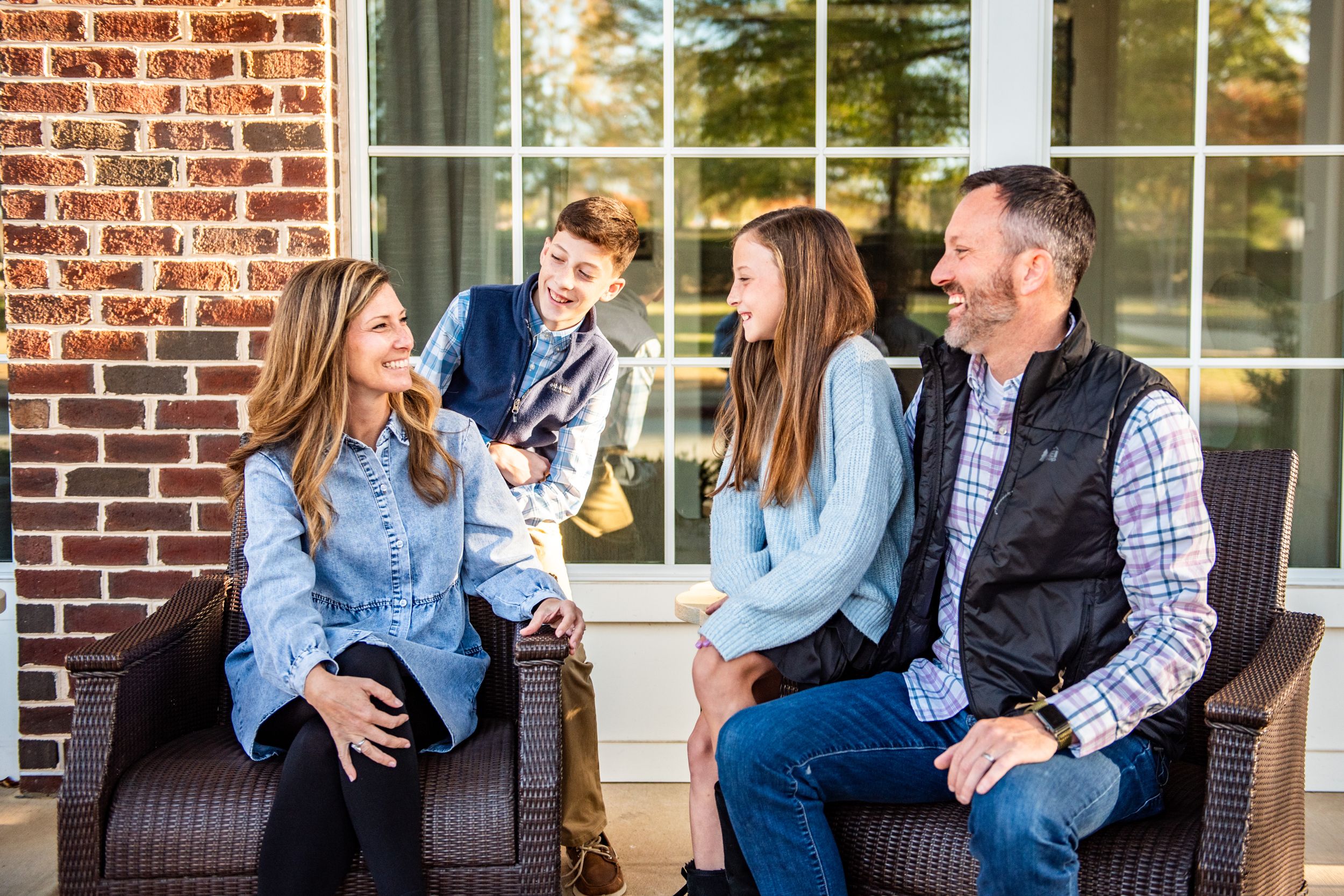 A family relaxes together on outdoor patio furniture in front of a brick home.