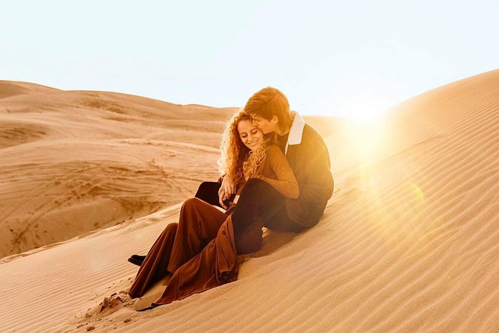 A person in dark clothing sits contemplatively on a sand dune during golden hour.