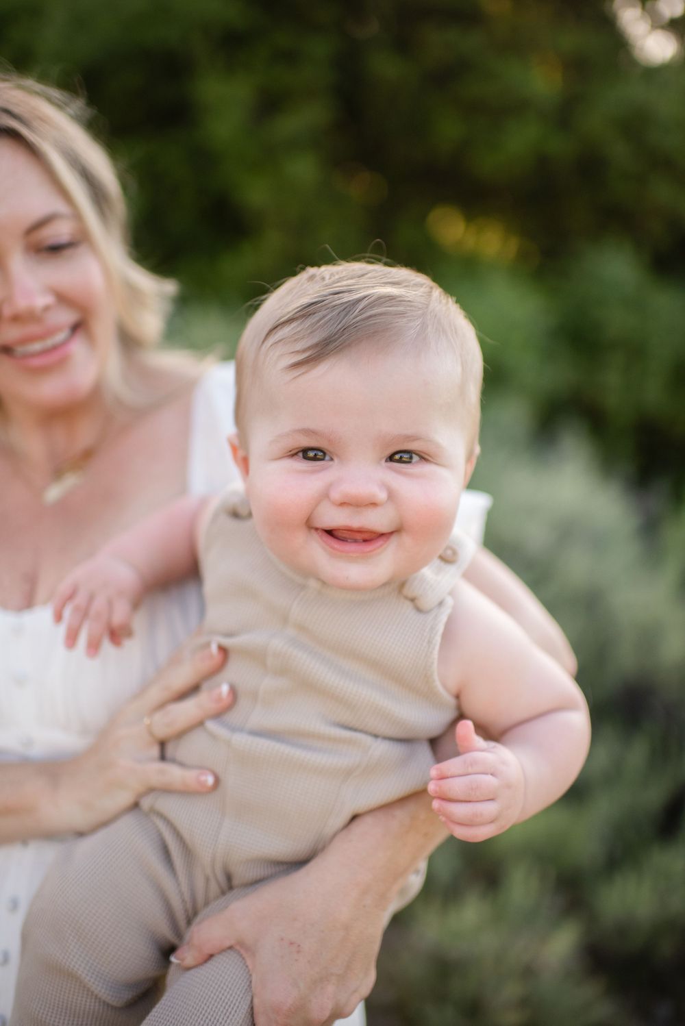 A baby in beige clothing smiles brightly while being held outdoors.