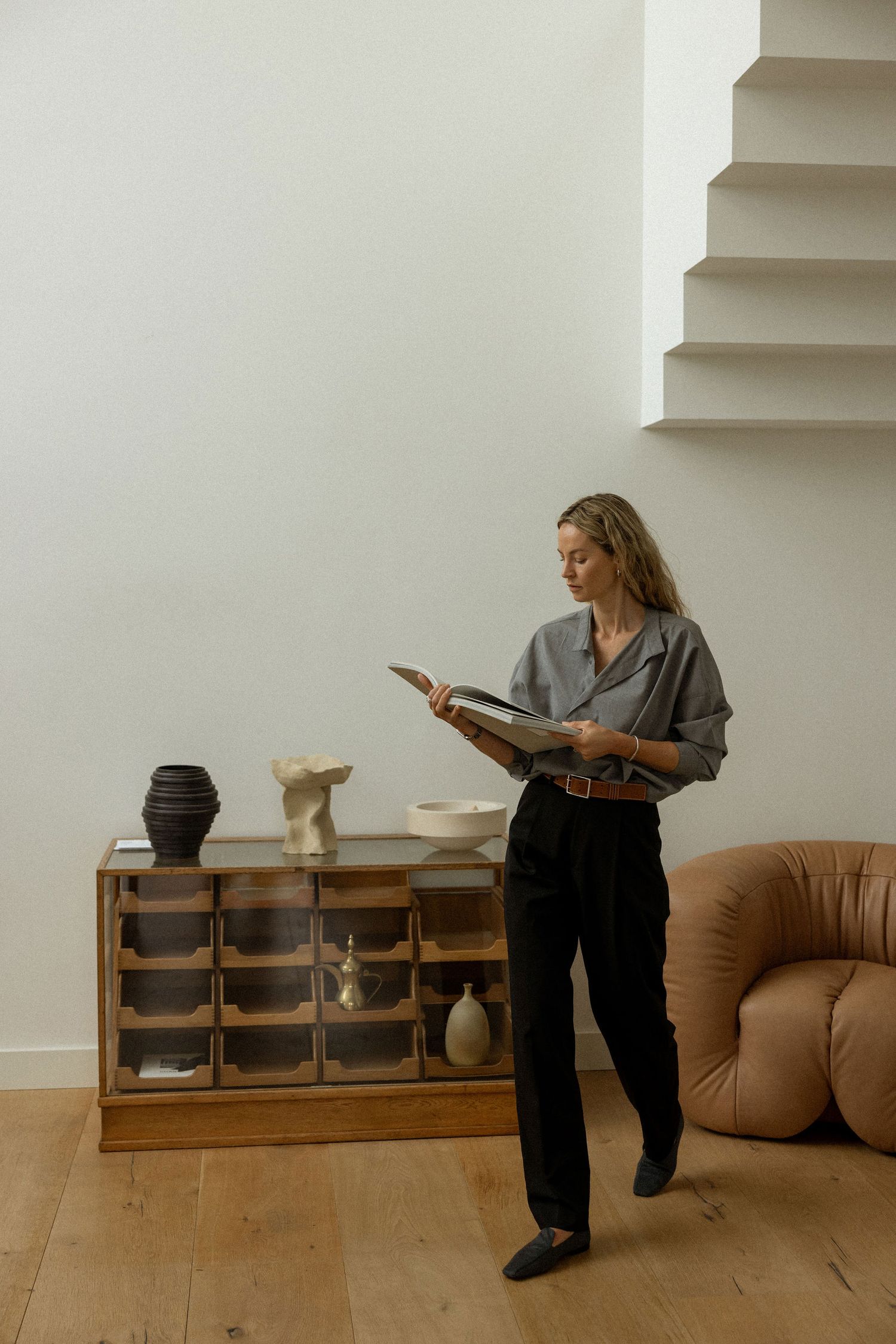 Woman reading book while standing in modern minimalist interior with wooden shelving.