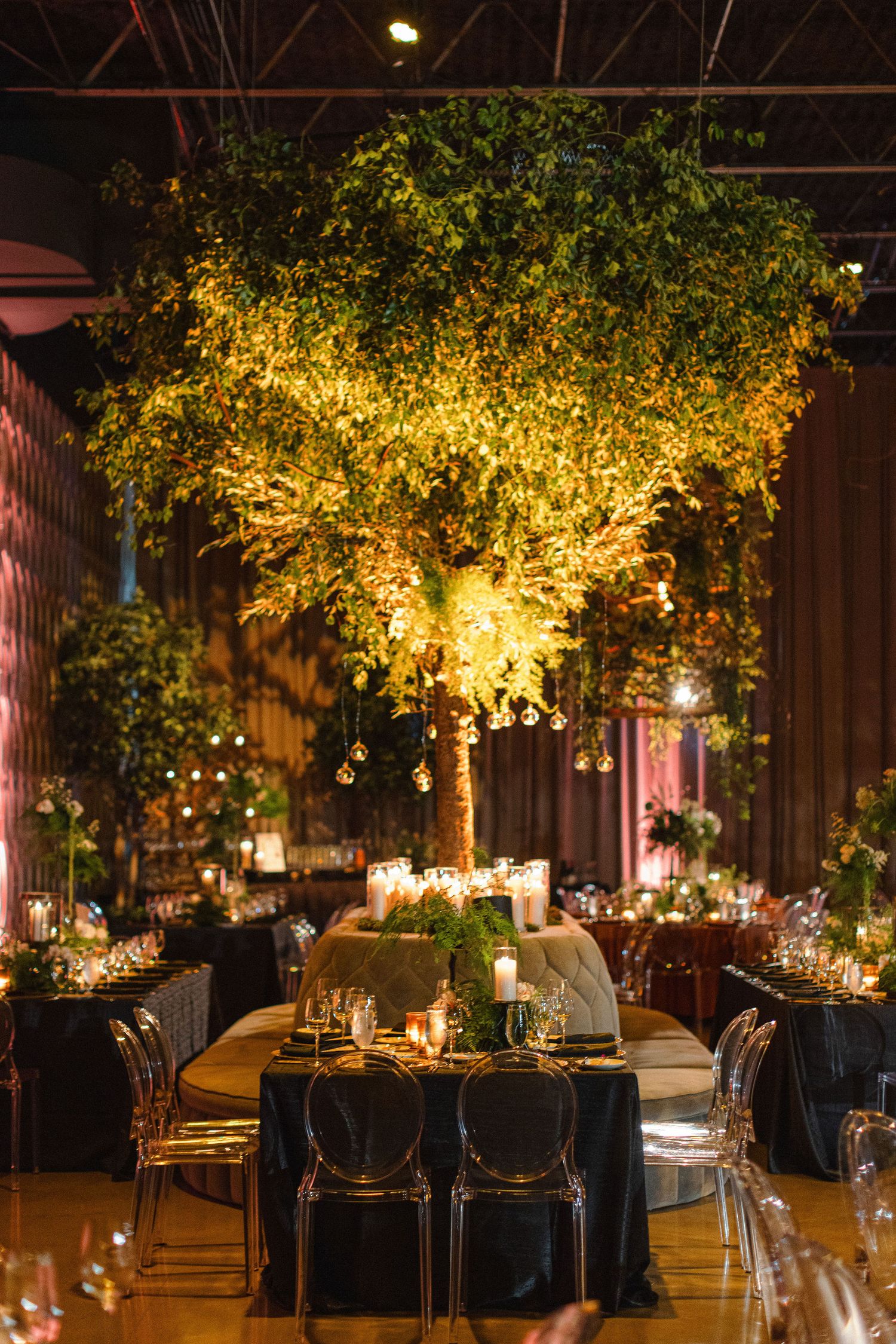 Elegant reception table setting under illuminated tree installation with black chairs and candlelight.