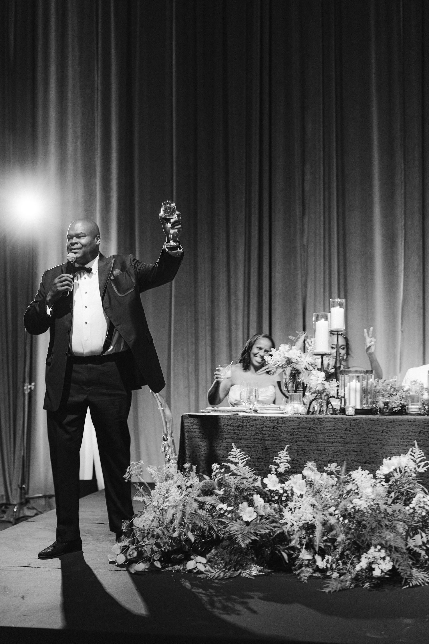 Wedding reception toast scene with dramatic stage lighting and floral decorations.