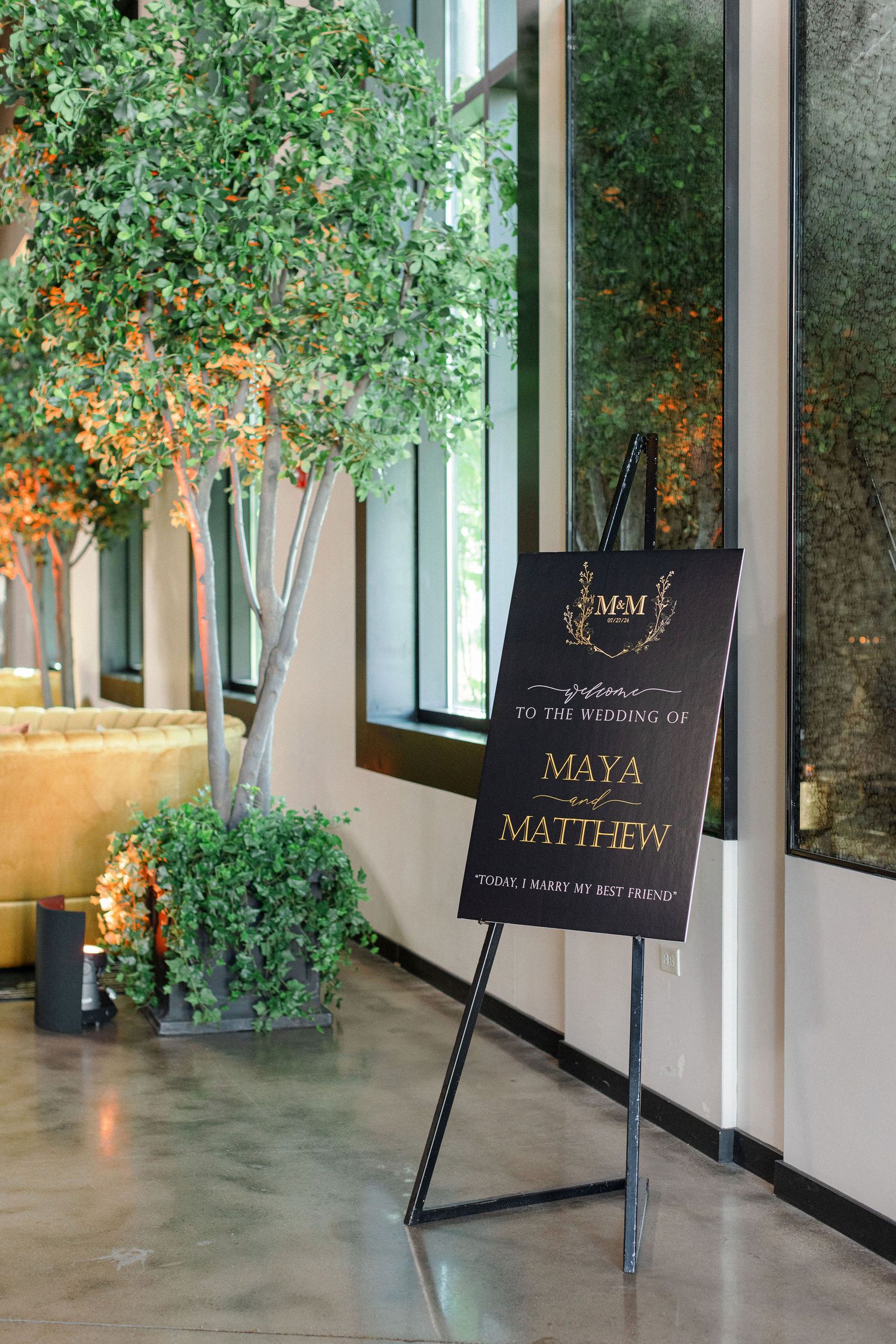 Elegant black and gold wedding welcome sign displayed with greenery in modern venue.