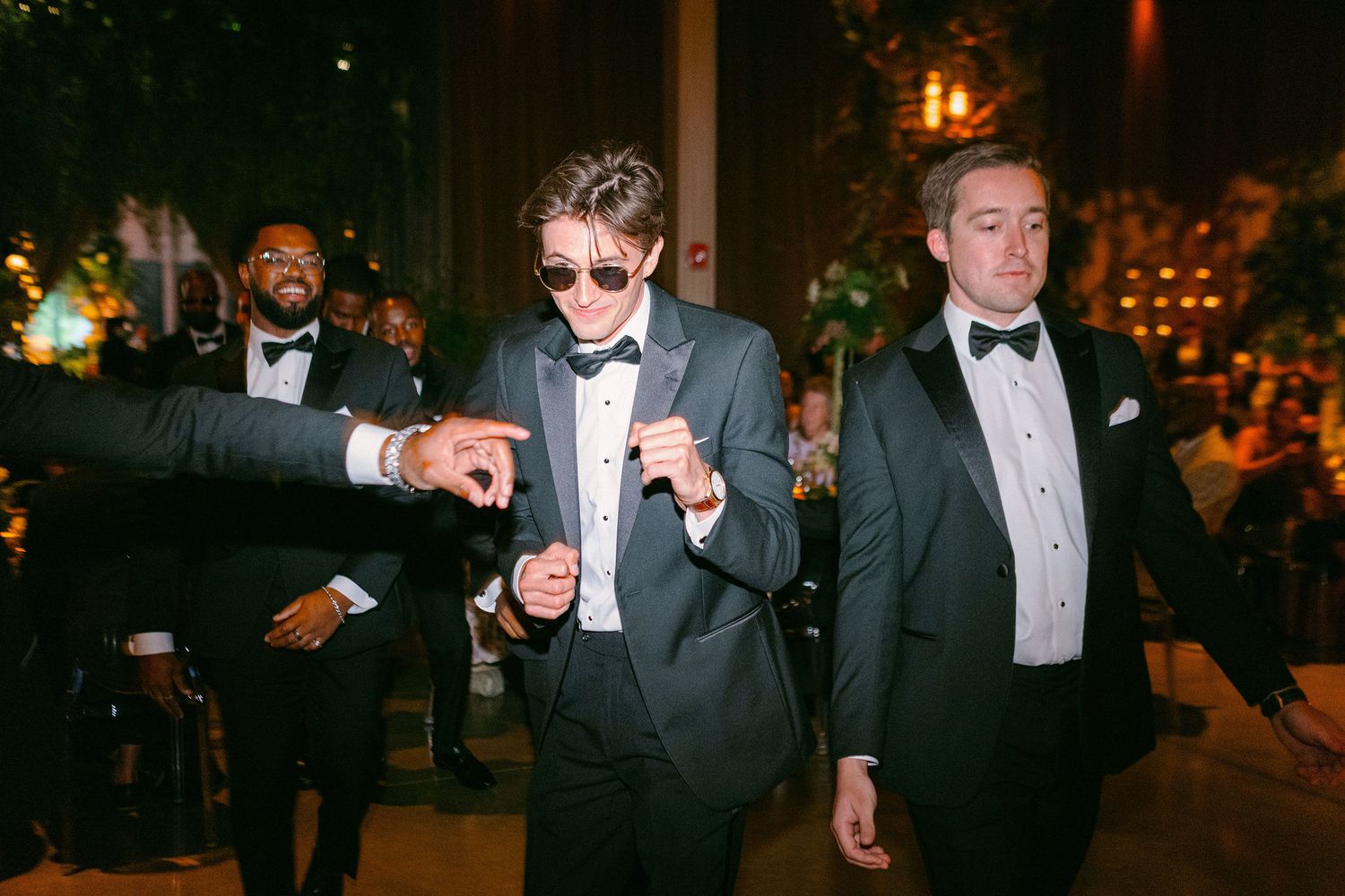 Three men in formal tuxedos walking through an elegant reception venue at night.