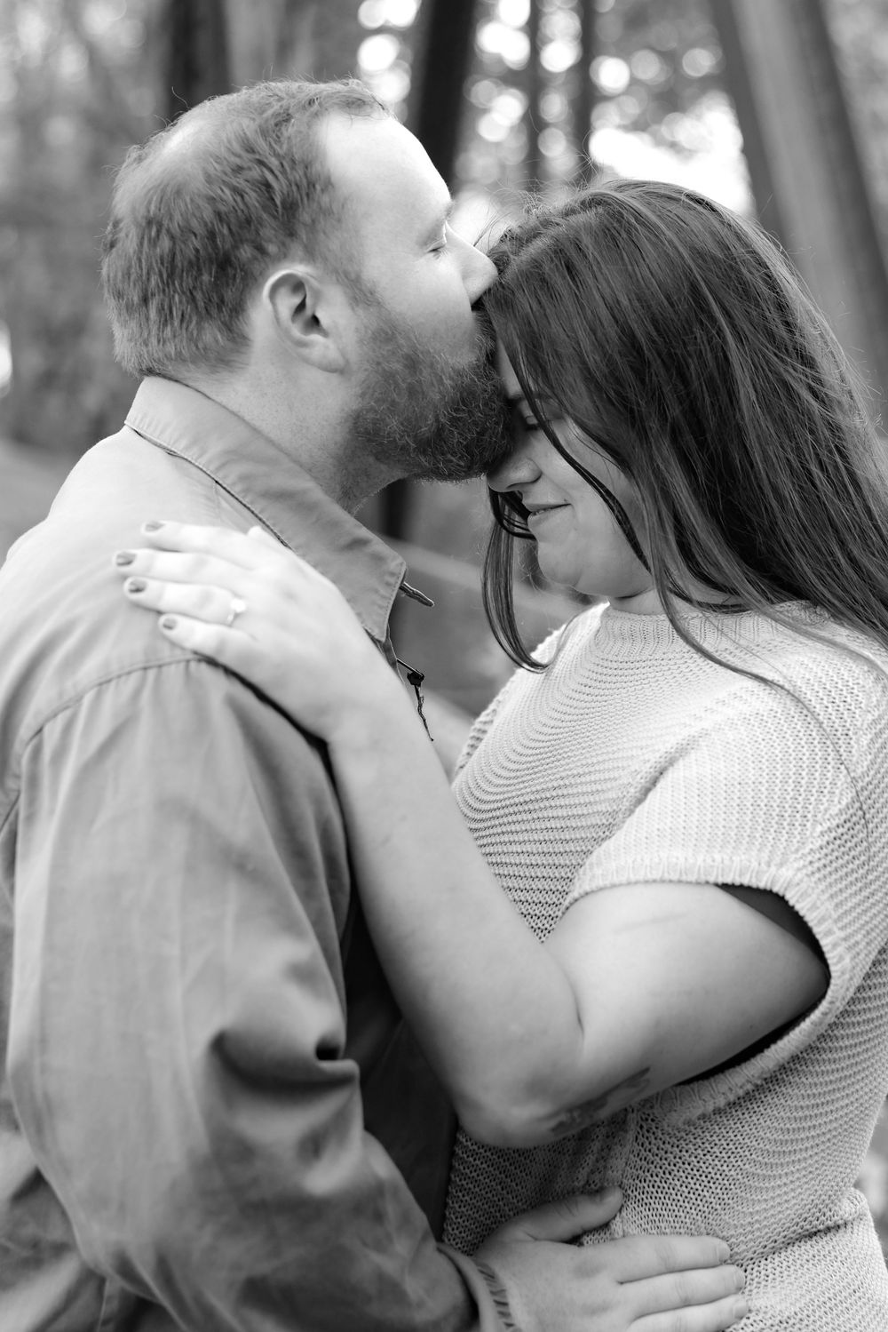Black and white romantic photo of bearded man kissing woman on forehead in forest setting.
