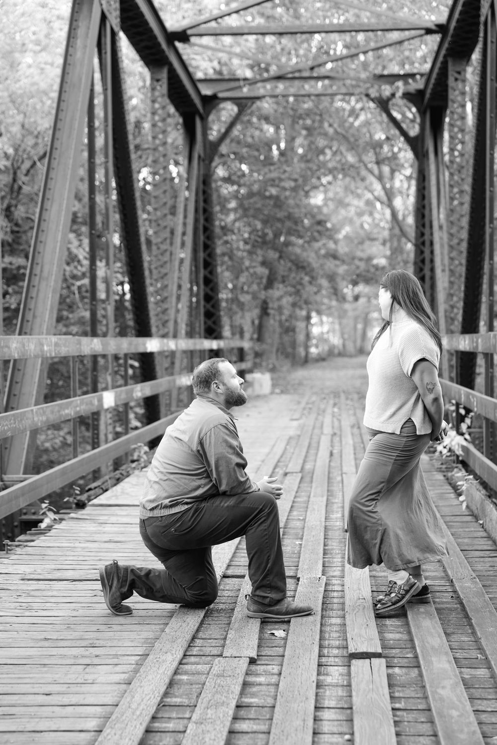 Black and white photo of man proposing on one knee to woman on a rustic wooden bridge.