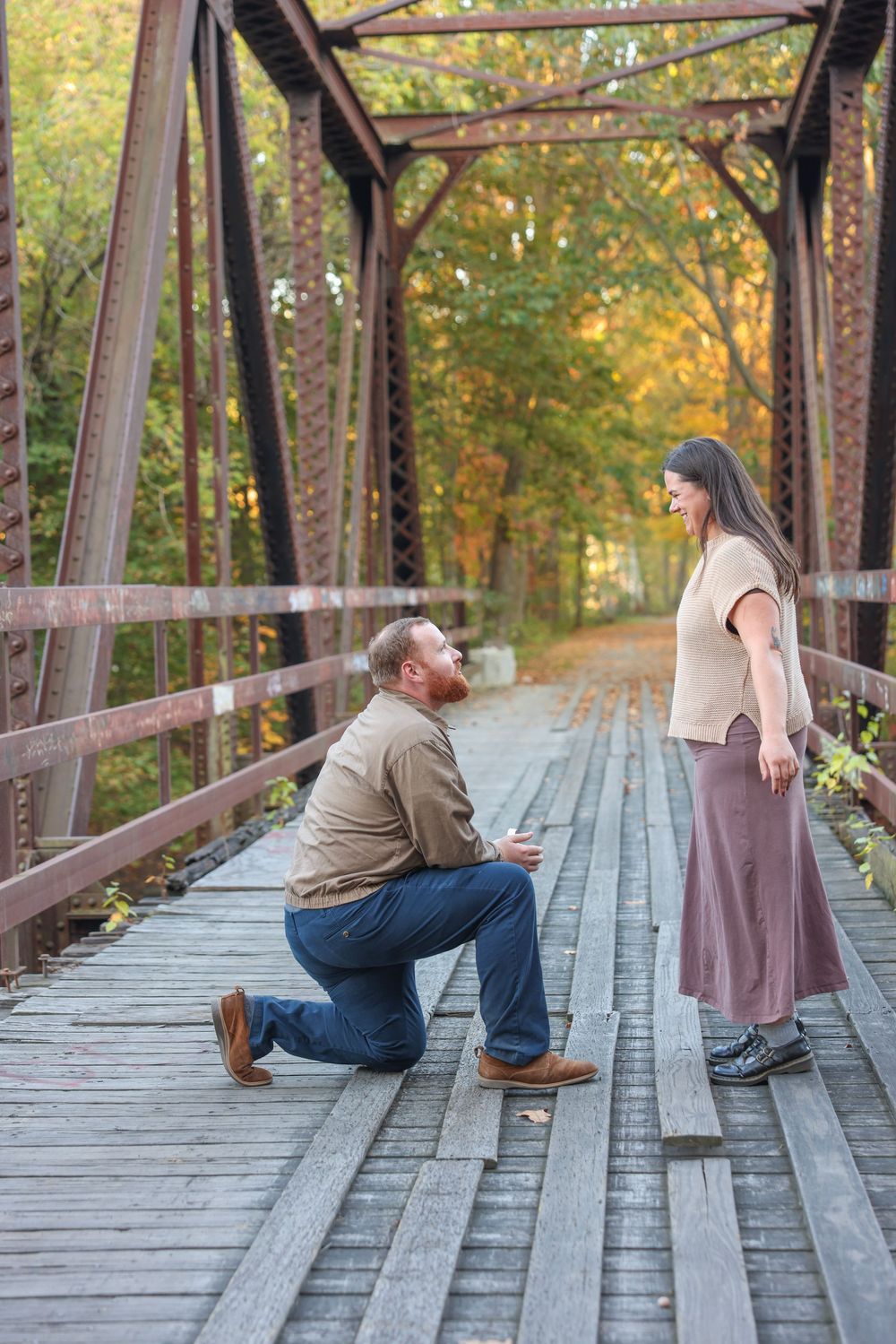 Man proposing on one knee to woman on a wooden bridge in fall.