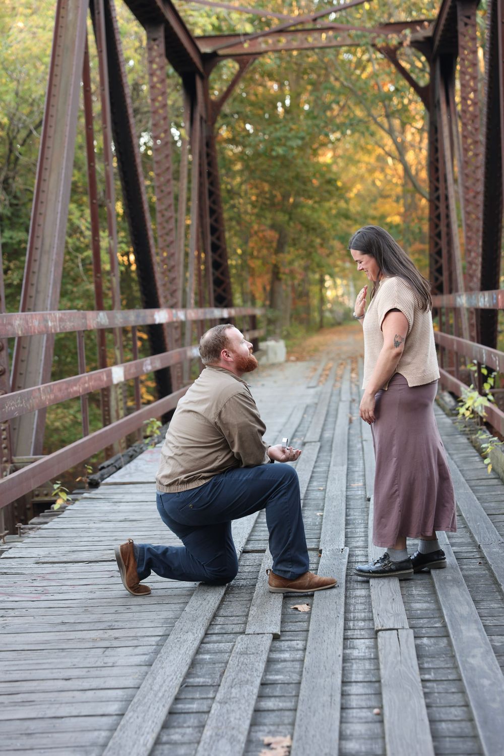 Man proposing on one knee to woman on a wooden iron bridge during fall engagement session.
