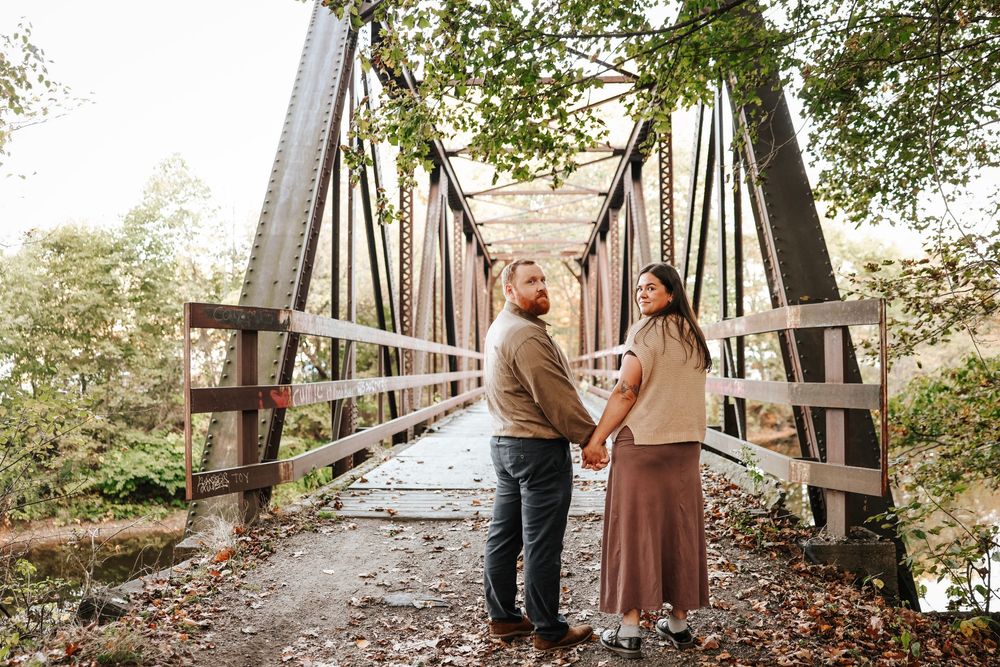 Couple holding hands on a rustic iron bridge surrounded by autumn foliage.