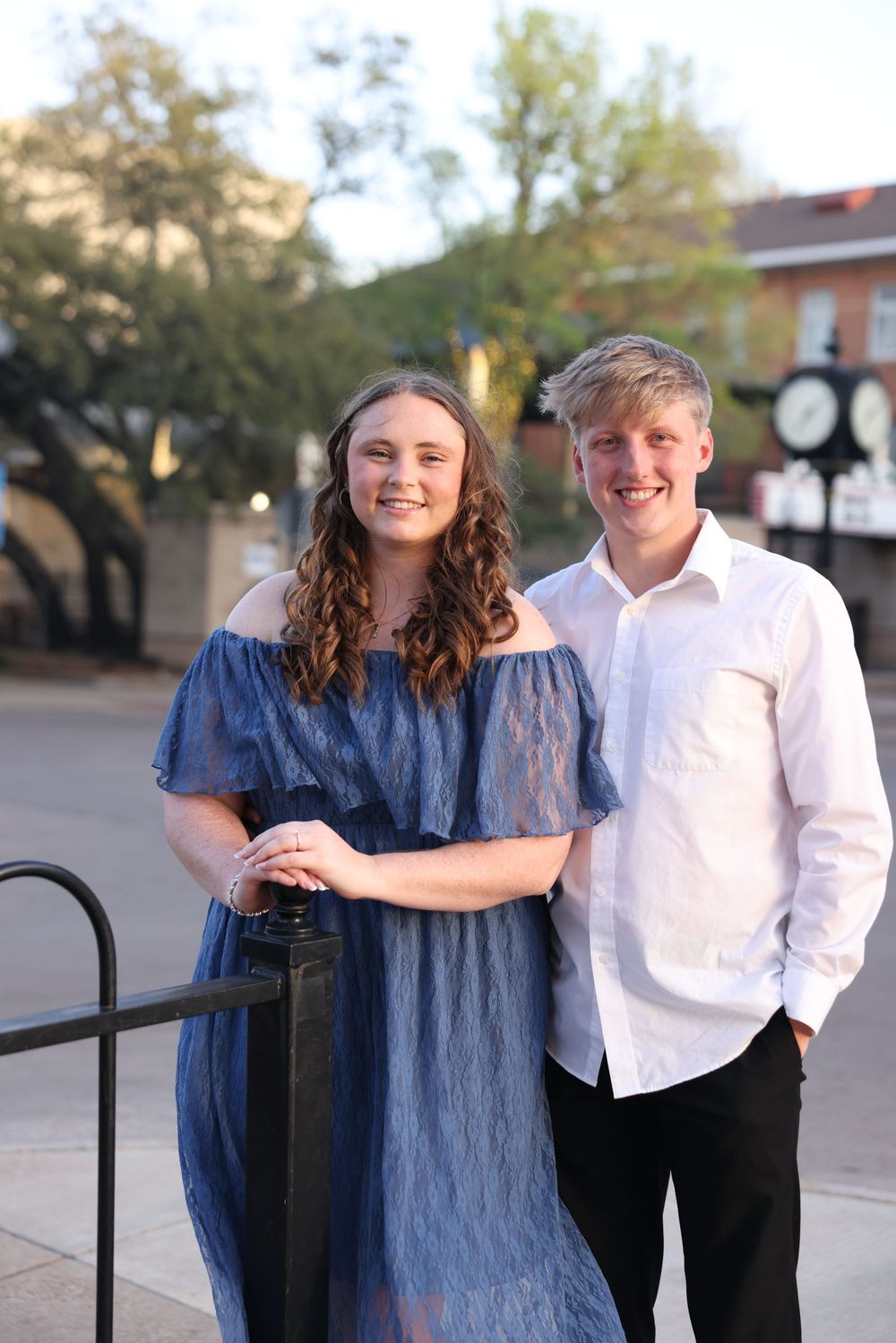 Smiling young couple posing outdoors at golden hour, woman in blue lace dress and man in white shirt.