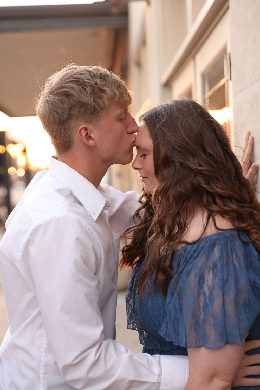 Young couple sharing a romantic forehead kiss against a warmly lit building at sunset.