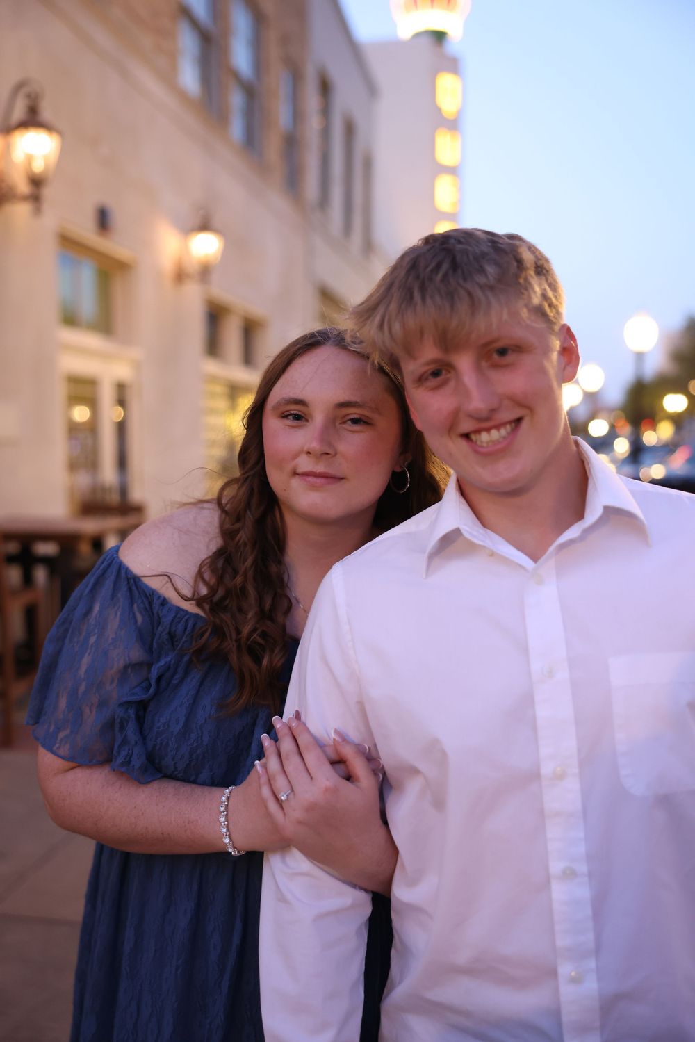 Young couple posing together at dusk on a city street, woman in blue dress and man in white shirt smiling.