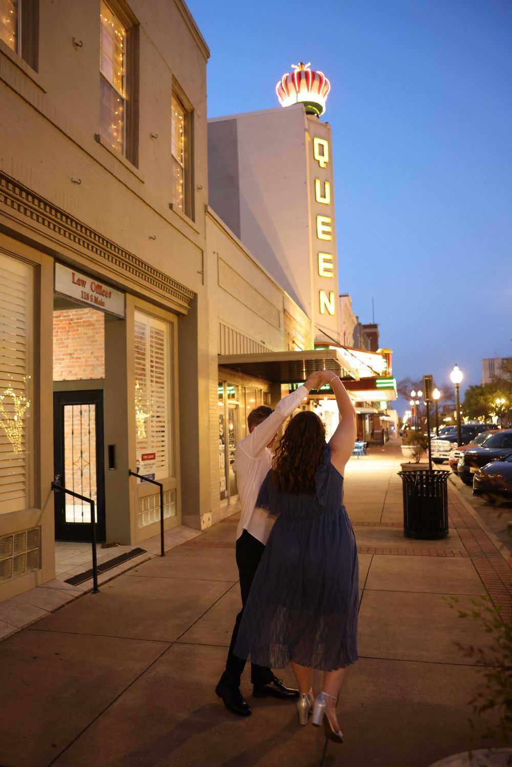 Couple dancing outside the Queen Theatre at night, captured from behind on a city sidewalk.
