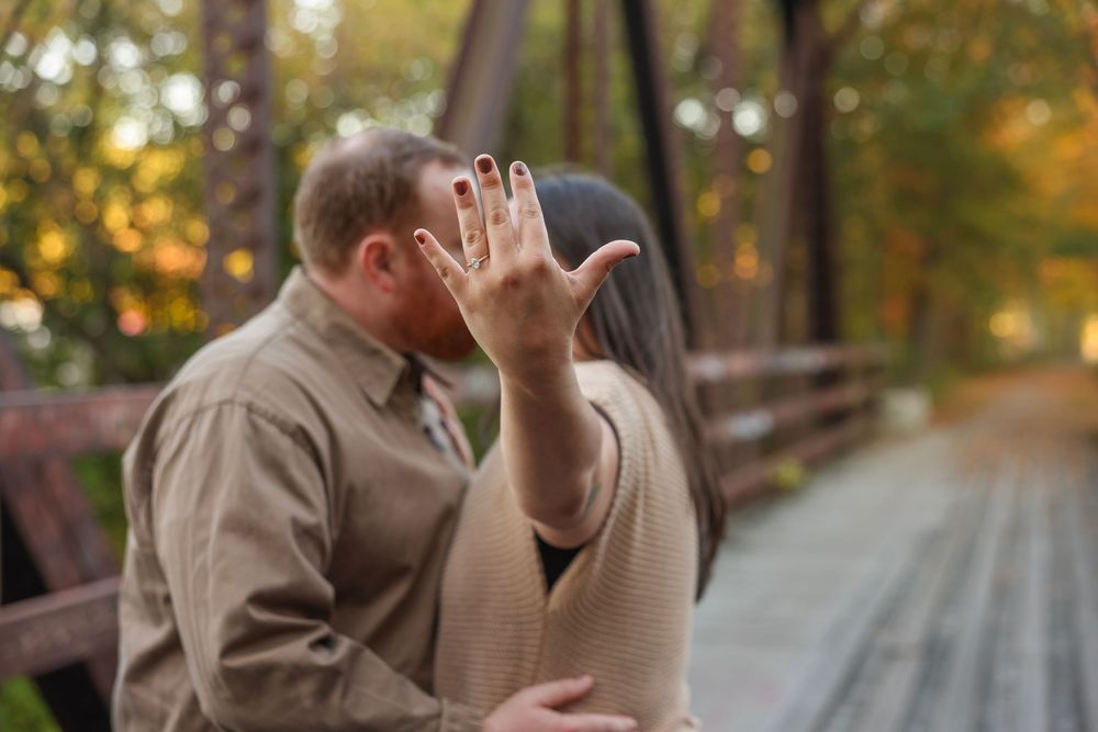 Engaged couple kissing on bridge while woman shows off diamond engagement ring to camera.