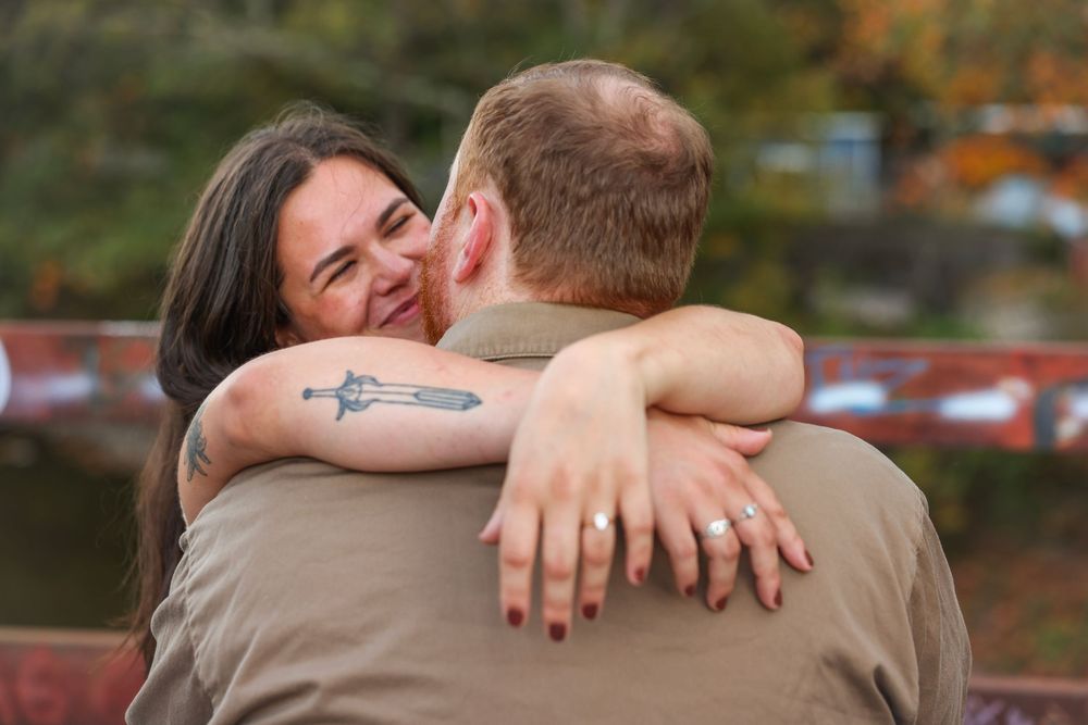 Woman with tattoo smiling and hugging man outdoors during fall engagement photo session.