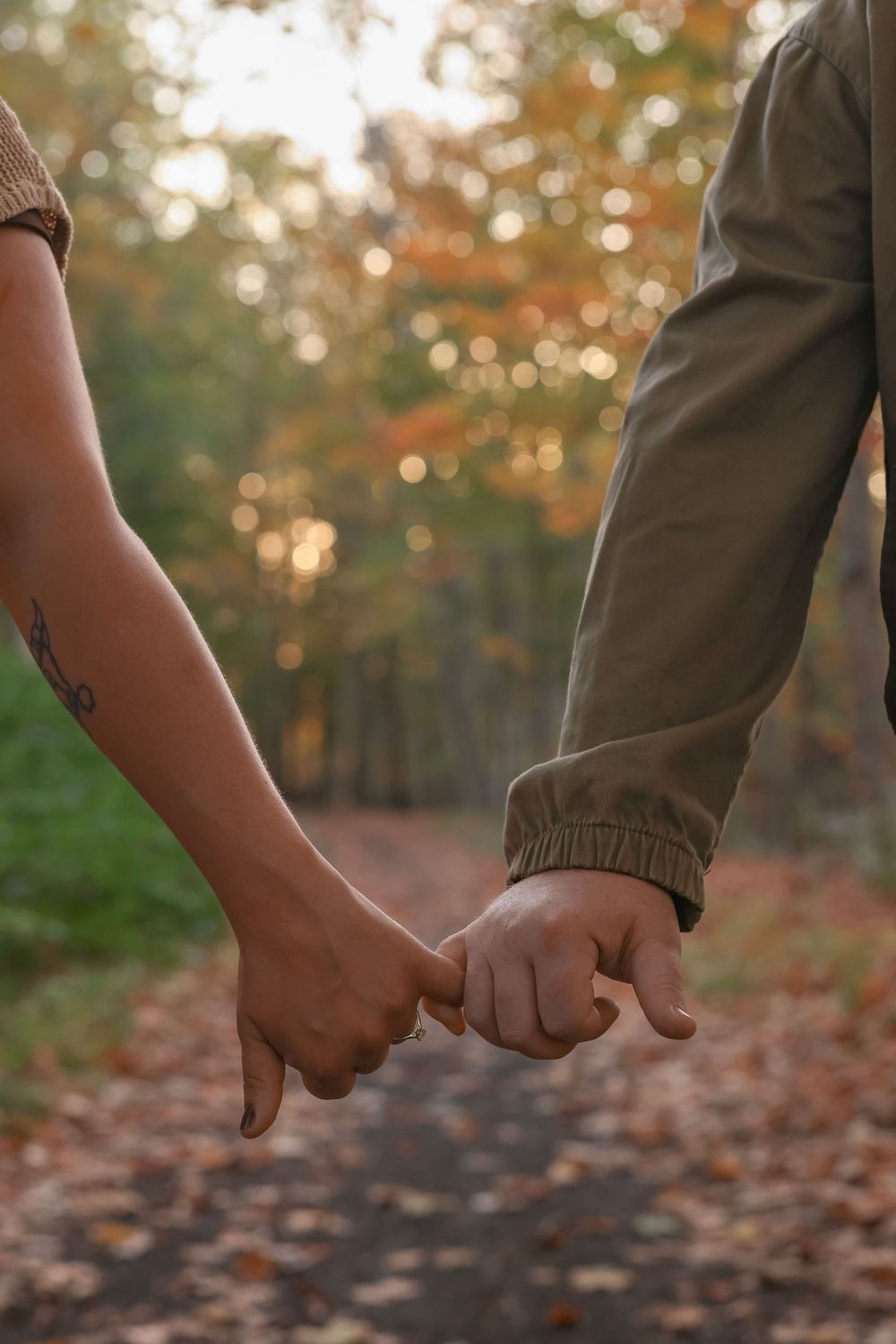 Close-up of couple holding pinky fingers on an autumn forest path with colorful fall foliage.