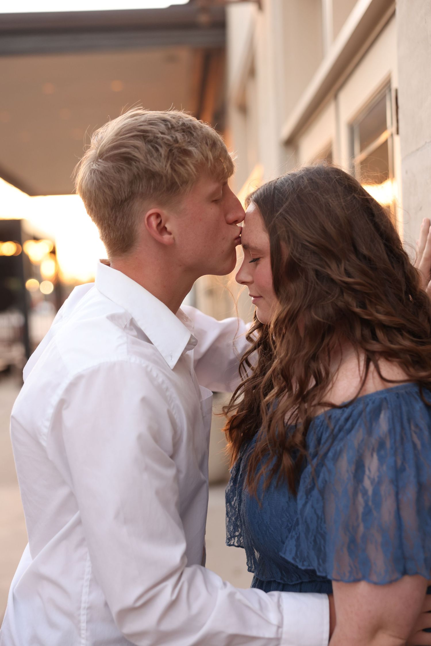 Young couple sharing a tender forehead kiss outdoors at golden hour during engagement session.