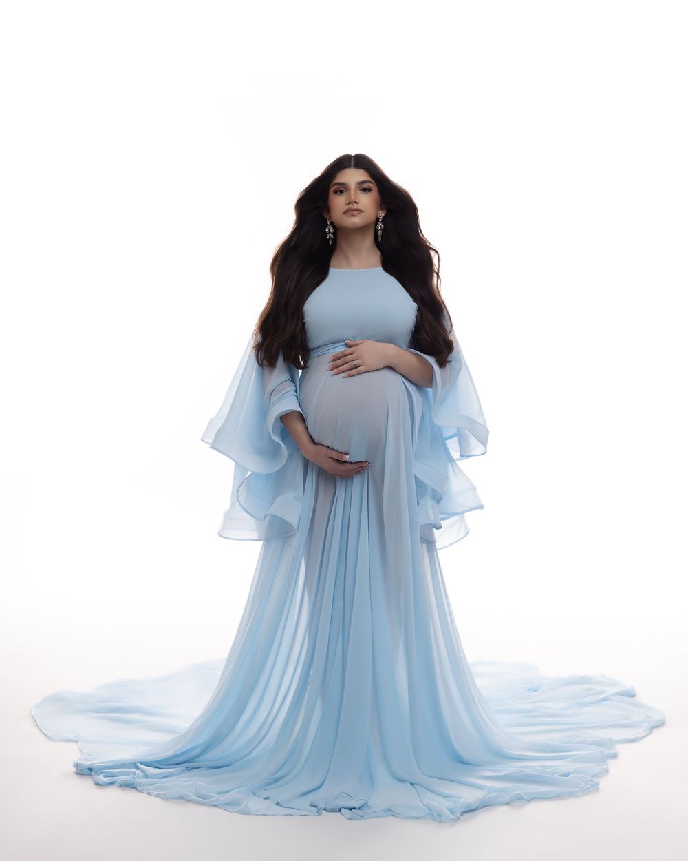 Pregnant woman in elegant light blue chiffon maternity gown with flowing sleeves and train, posing against white background.