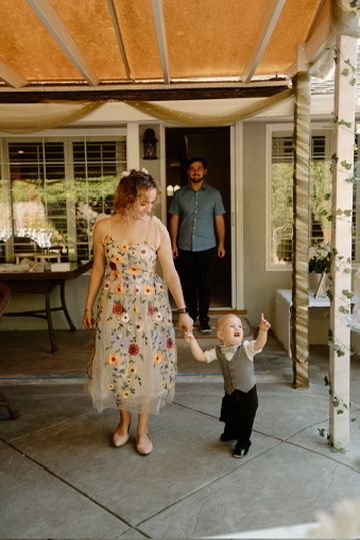 A family spends time together on a covered porch, with a woman in a floral dress walking with a toddler.