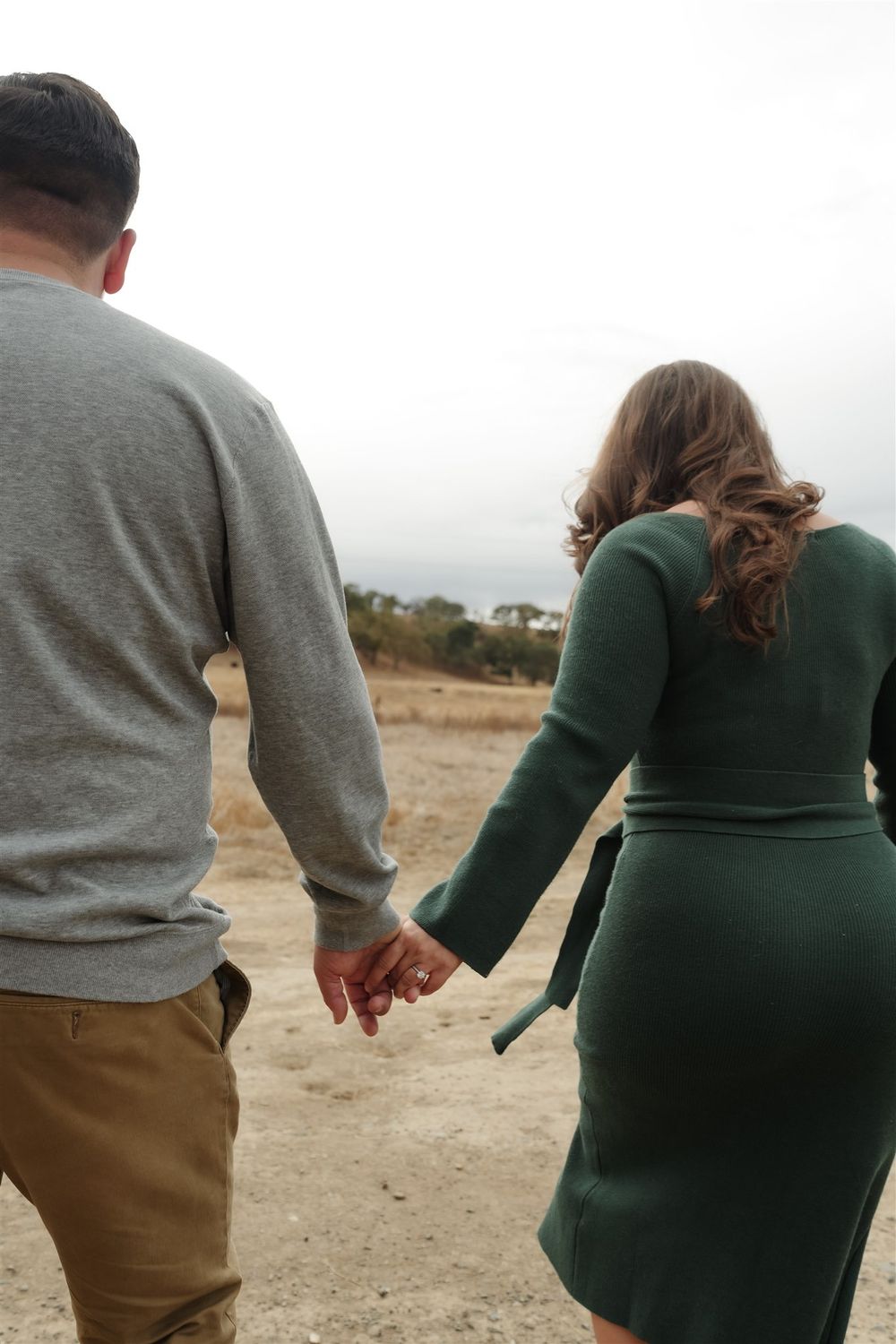 A couple holds hands while walking on a grass path, with person in gray sweater and another in green dress.