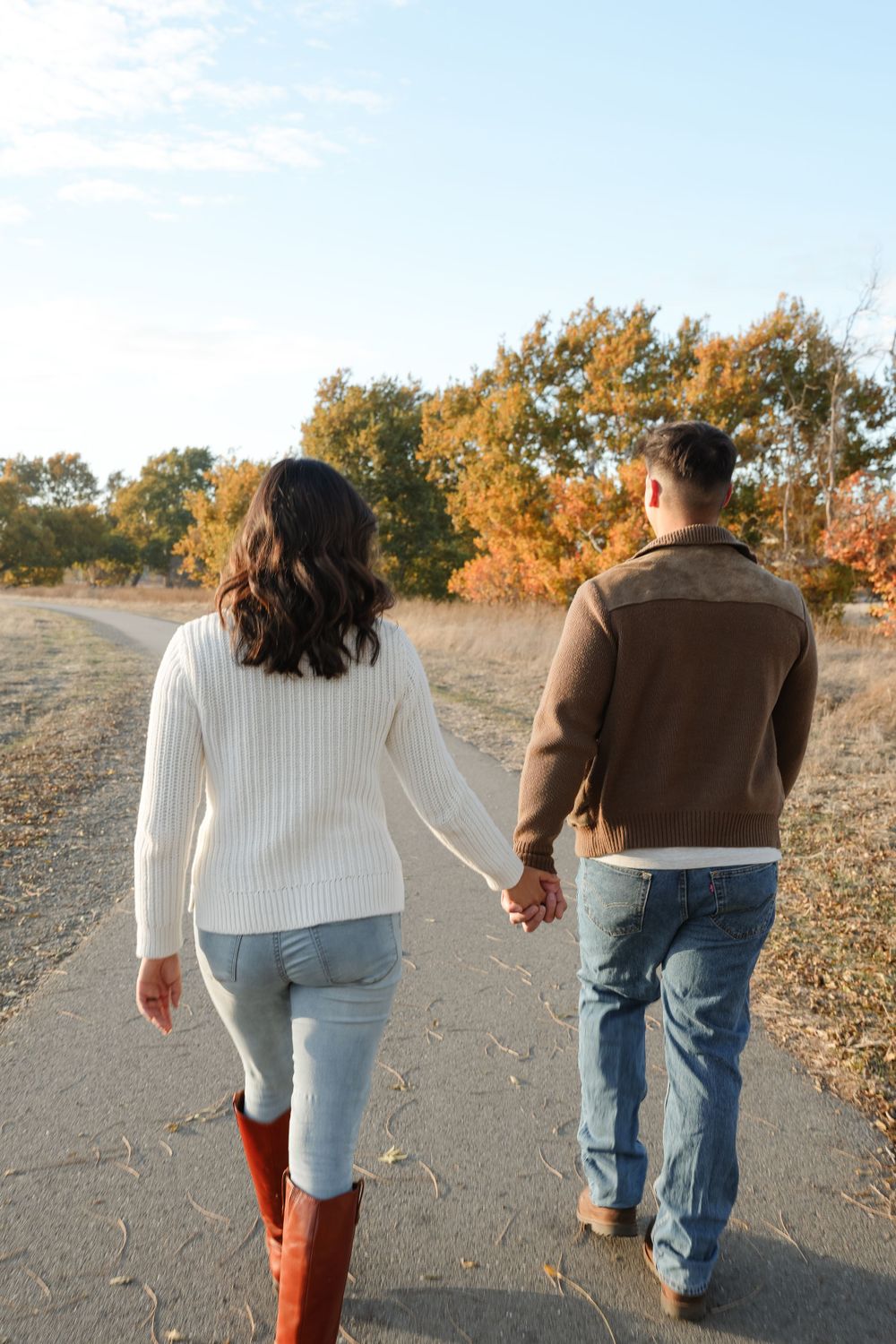 A couple holds hands while walking together on a path surrounded by autumn trees at golden hour.