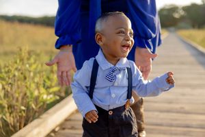 An adorable toddler boy in suspenders and bow tie laughs joyfully on a wooden boardwalk in Matawan, NJ.