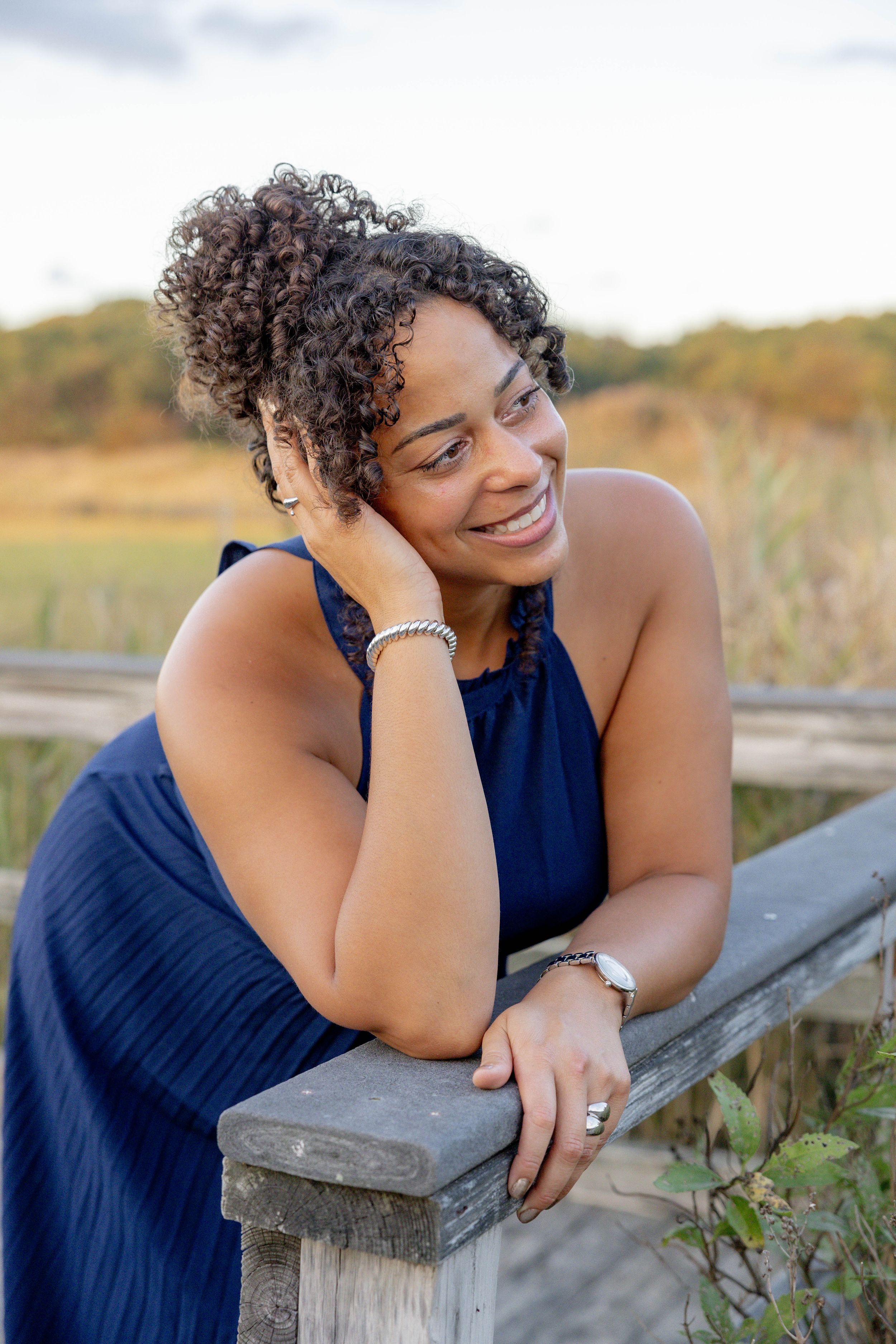 A smiling woman in a navy dress leans on a wooden fence railing outdoors in Matawan, NJ.