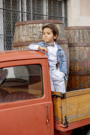 A stylish young boy in a bow tie and suspenders stands in a vintage red pickup truck bed in the Trastevere neighborhood of Rome, Italy.