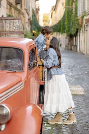 A mother kisses her young son standing in a vintage red truck on a charming cobblestone street in the Trastevere neighborhood of Rome, Italy