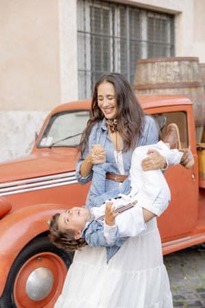 A laughing mother playfully lifts her young son outdoors near a vintage red classic car in the Trastevere neighborhood of Rome, Italy.