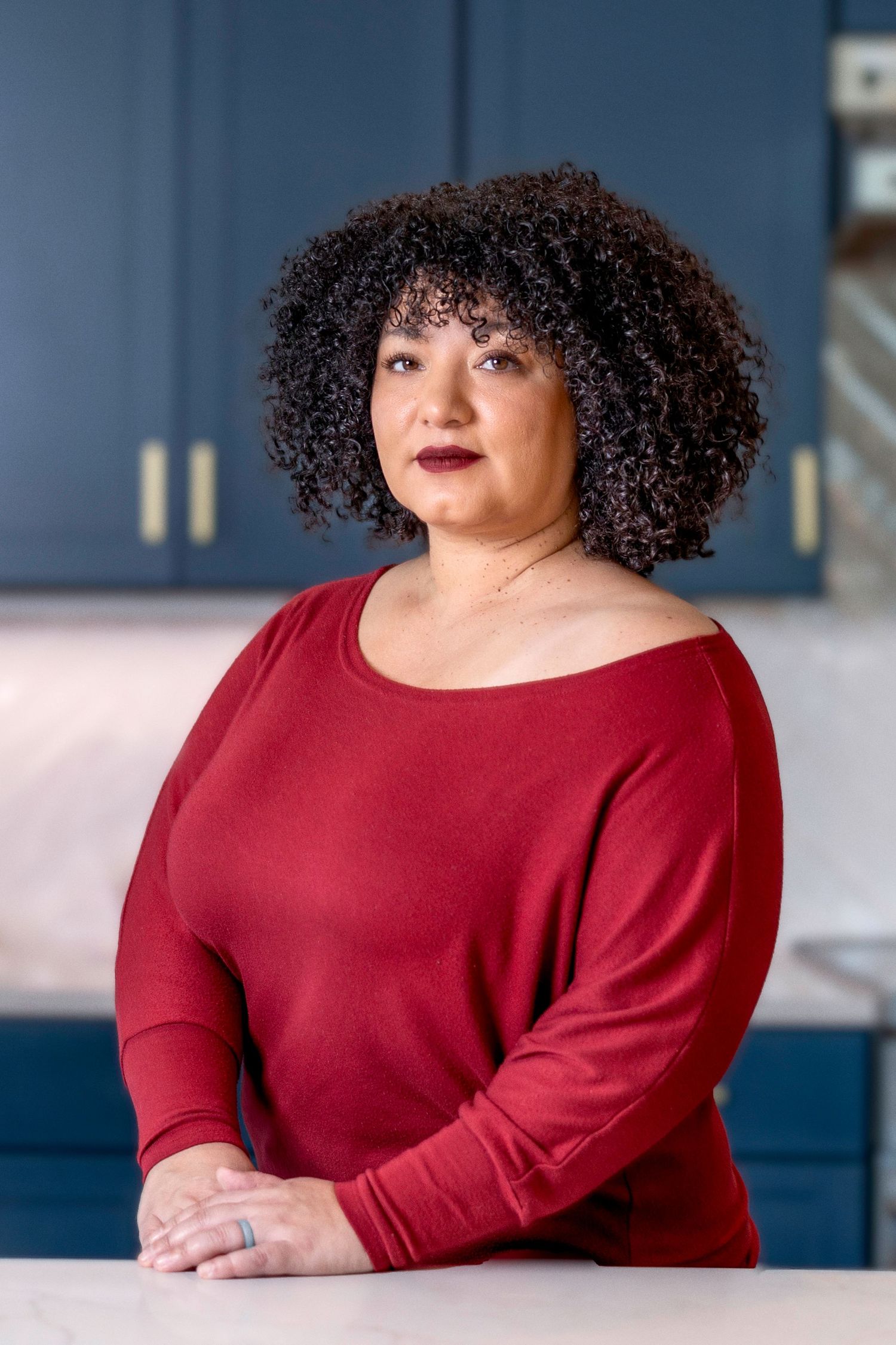 Person wearing a red long sleeve top poses against a blue wall with curly natural hair and deep red lipstick.