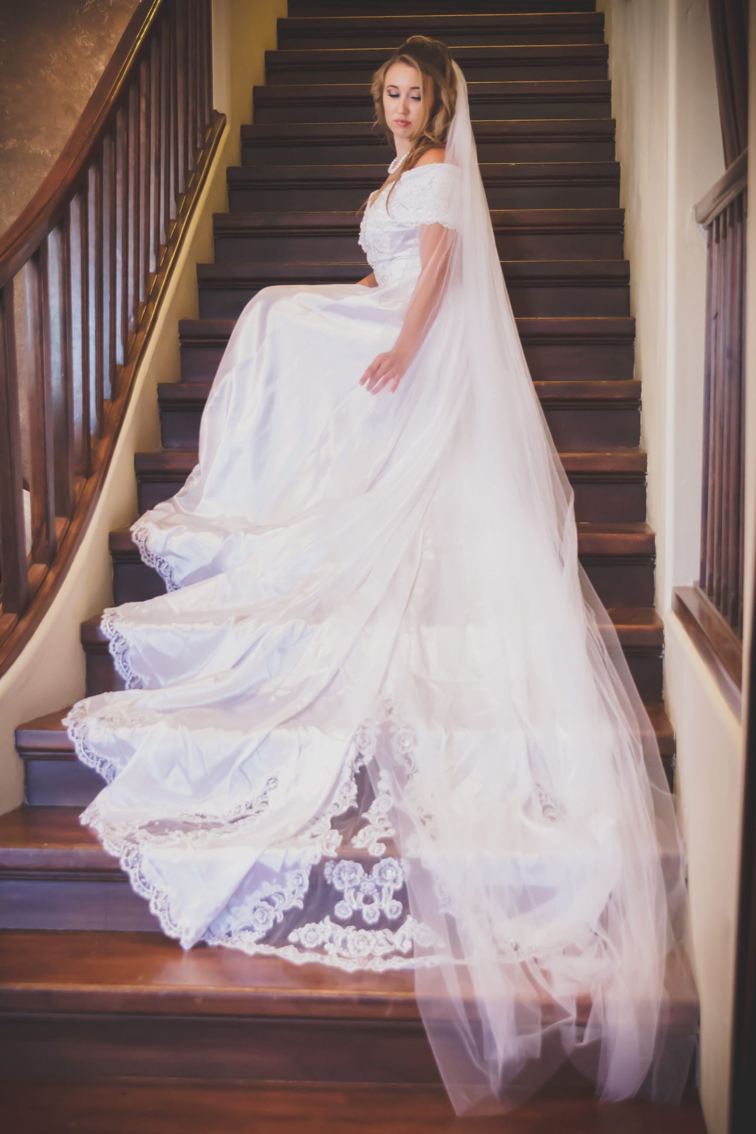 A bride in an elegant white gown with a flowing lace train and veil poses gracefully on a wooden staircase.